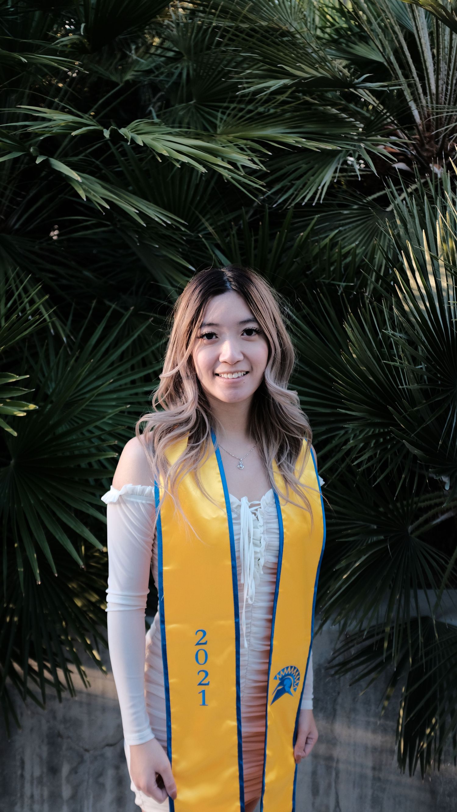 Graduate wearing yellow academic stole poses against palm frond background.
