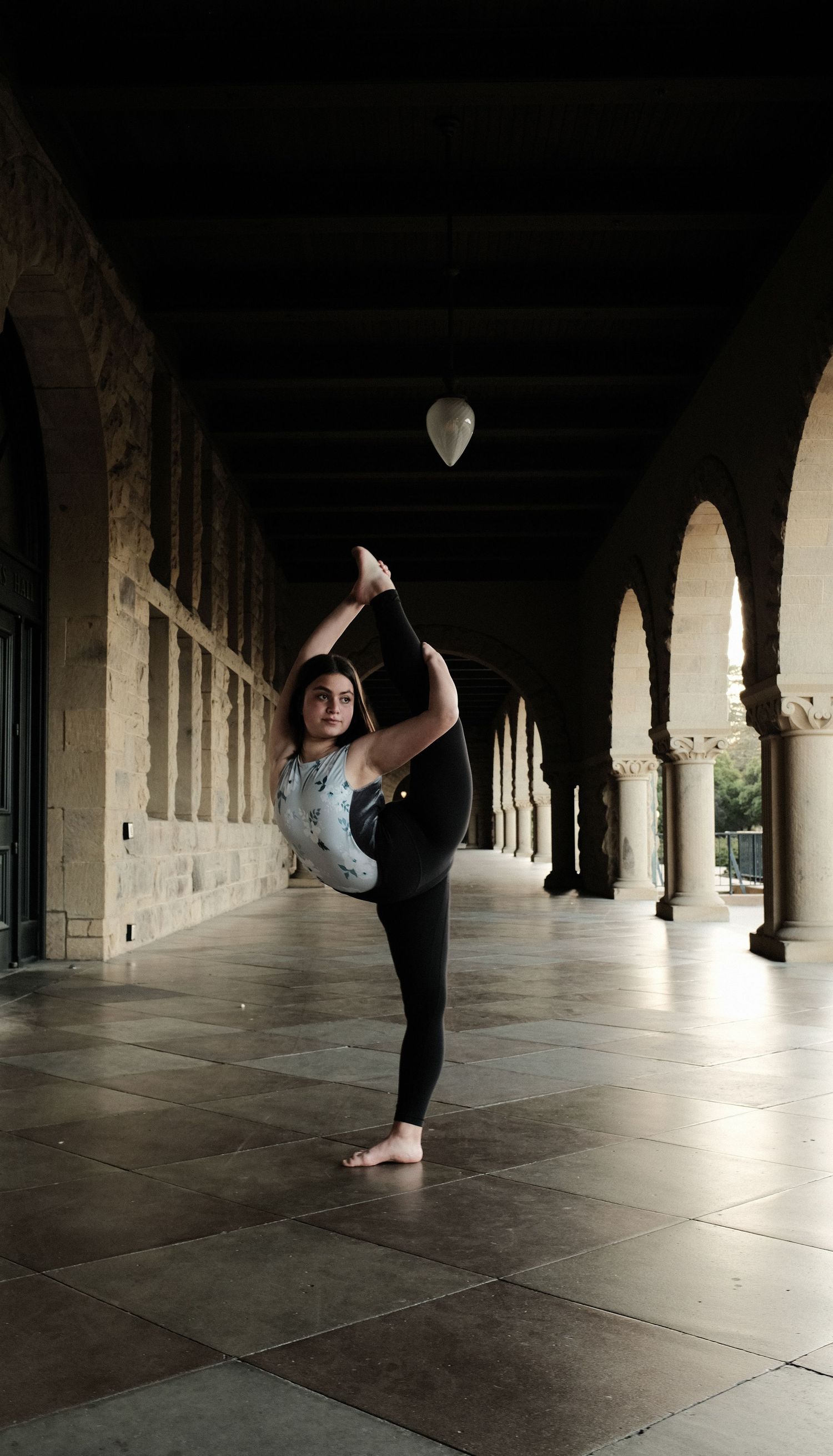 A dancer executes a standing split pose in a historic colonnade with arched architecture.