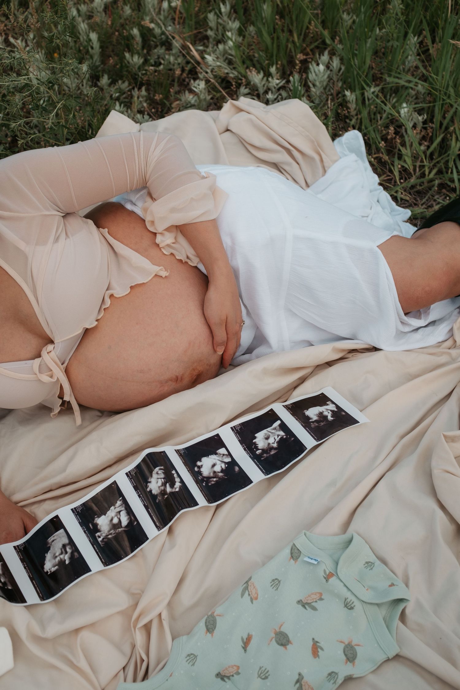 A pregnancy photo featuring ultrasound images and a mint onesie laying on a light blanket outdoors.