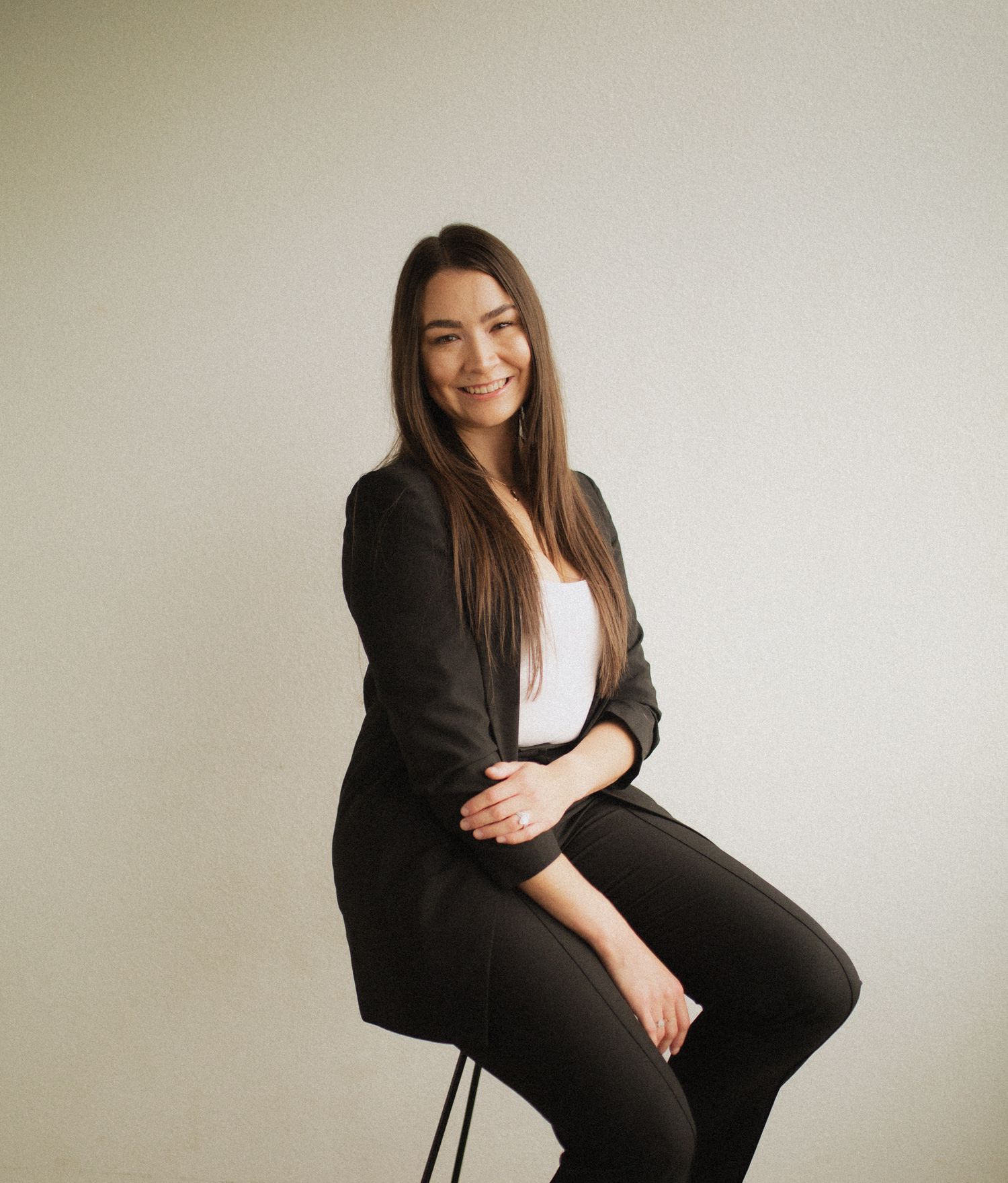 Woman in black business suit sits confidently in professional photoshoot against light backdrop.