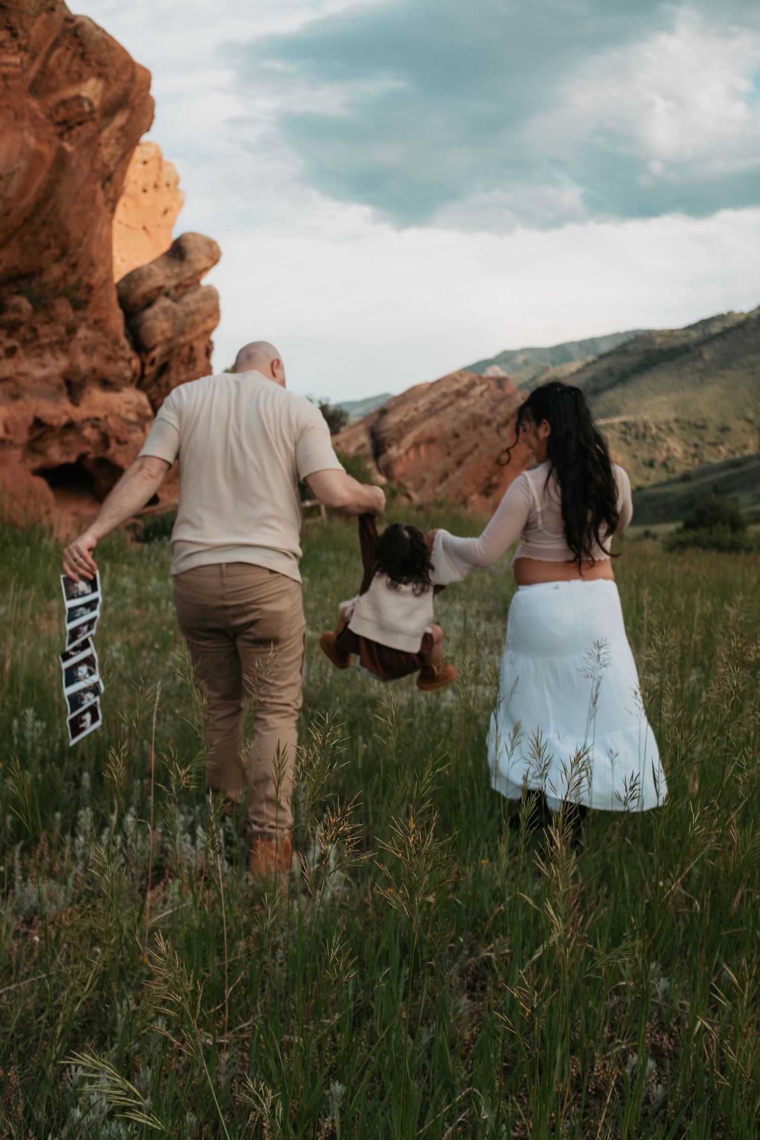 Family walking together through tall grass near dramatic red rock formations at sunset.