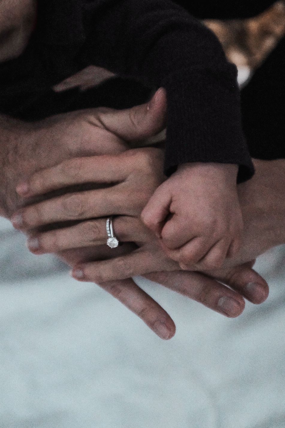 Close-up of intertwined hands with engagement ring in soft, moody lighting.
