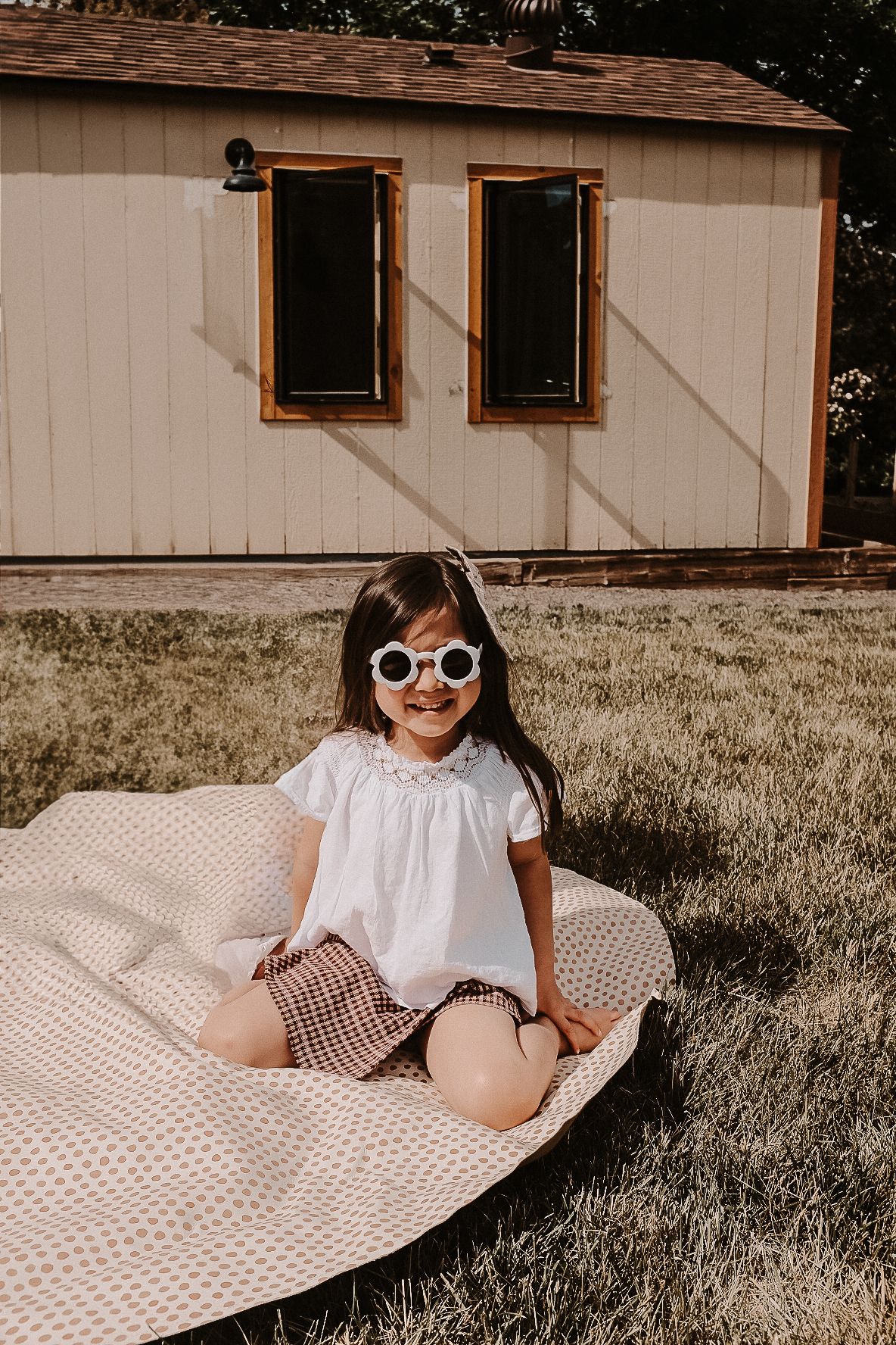 A young child wearing sunglasses sits on a striped blanket in front of a beige building.