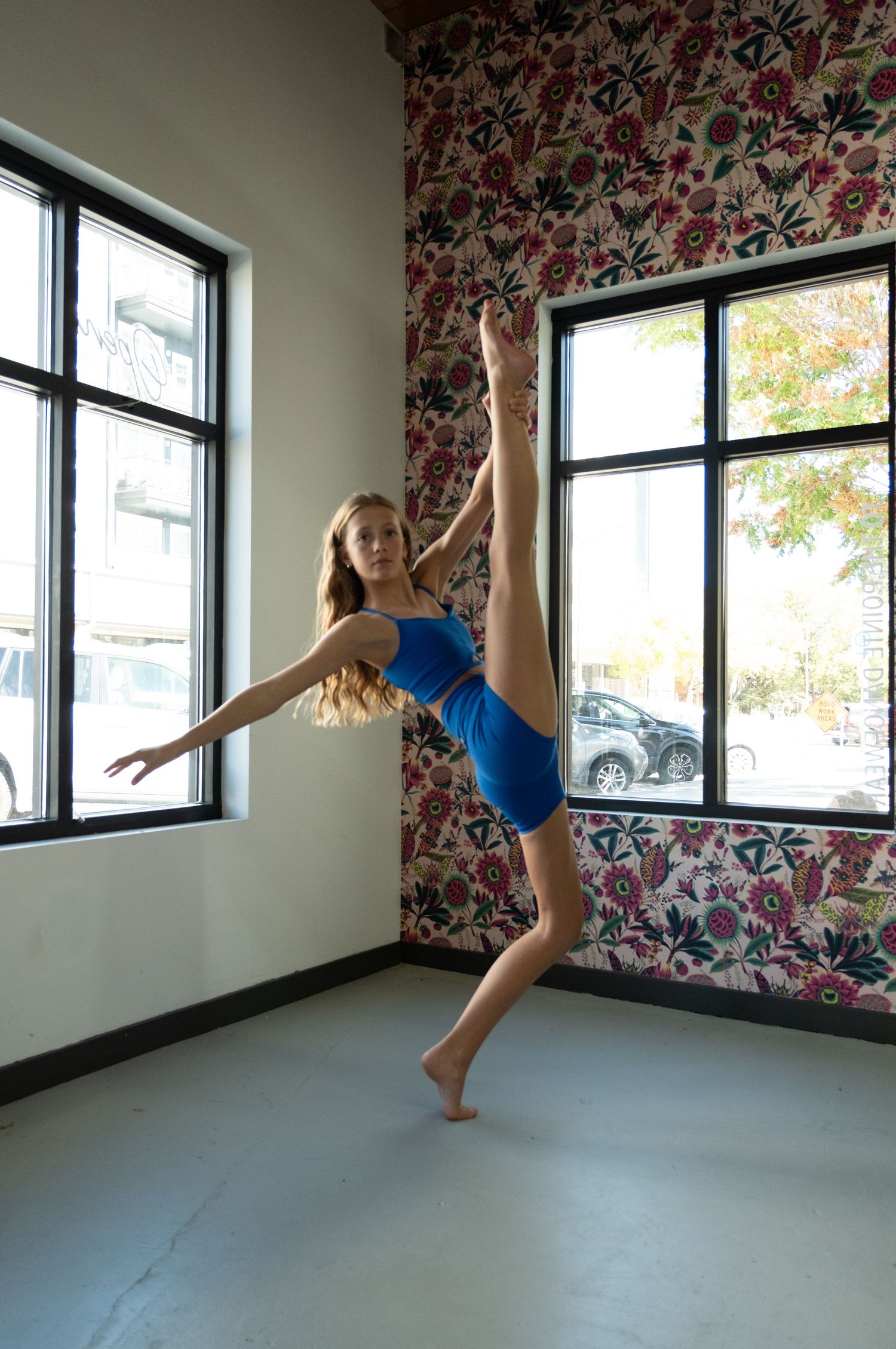 A flexible dancer in a blue outfit performs a high leg extension in a studio with floral wallpaper and large windows.