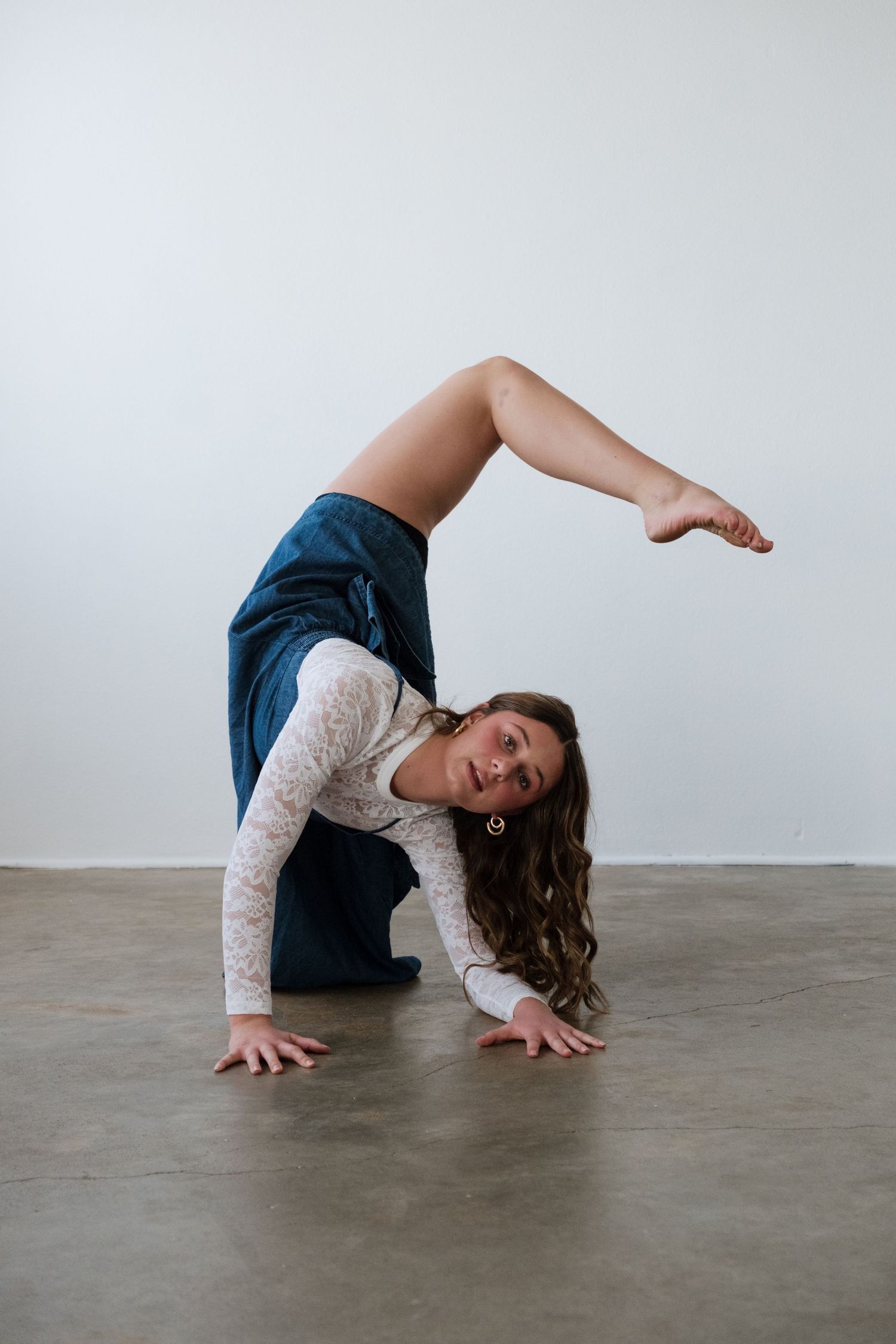Flexible dancer performs a backbend pose on concrete floor, wearing white lace top and teal skirt.