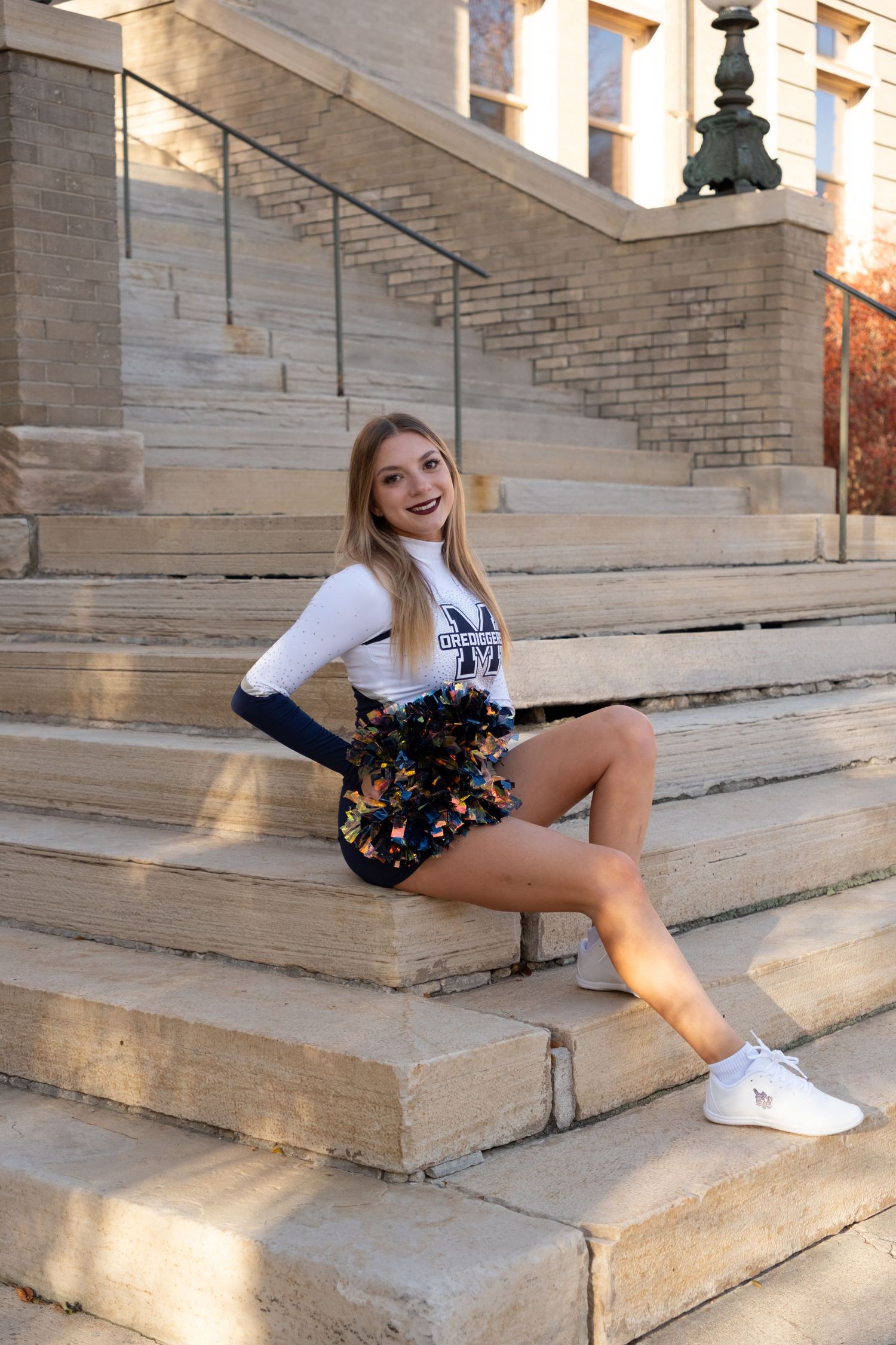 Smiling cheerleader in uniform sits on stone steps outdoors, wearing white top, dark skirt, and white sneakers.