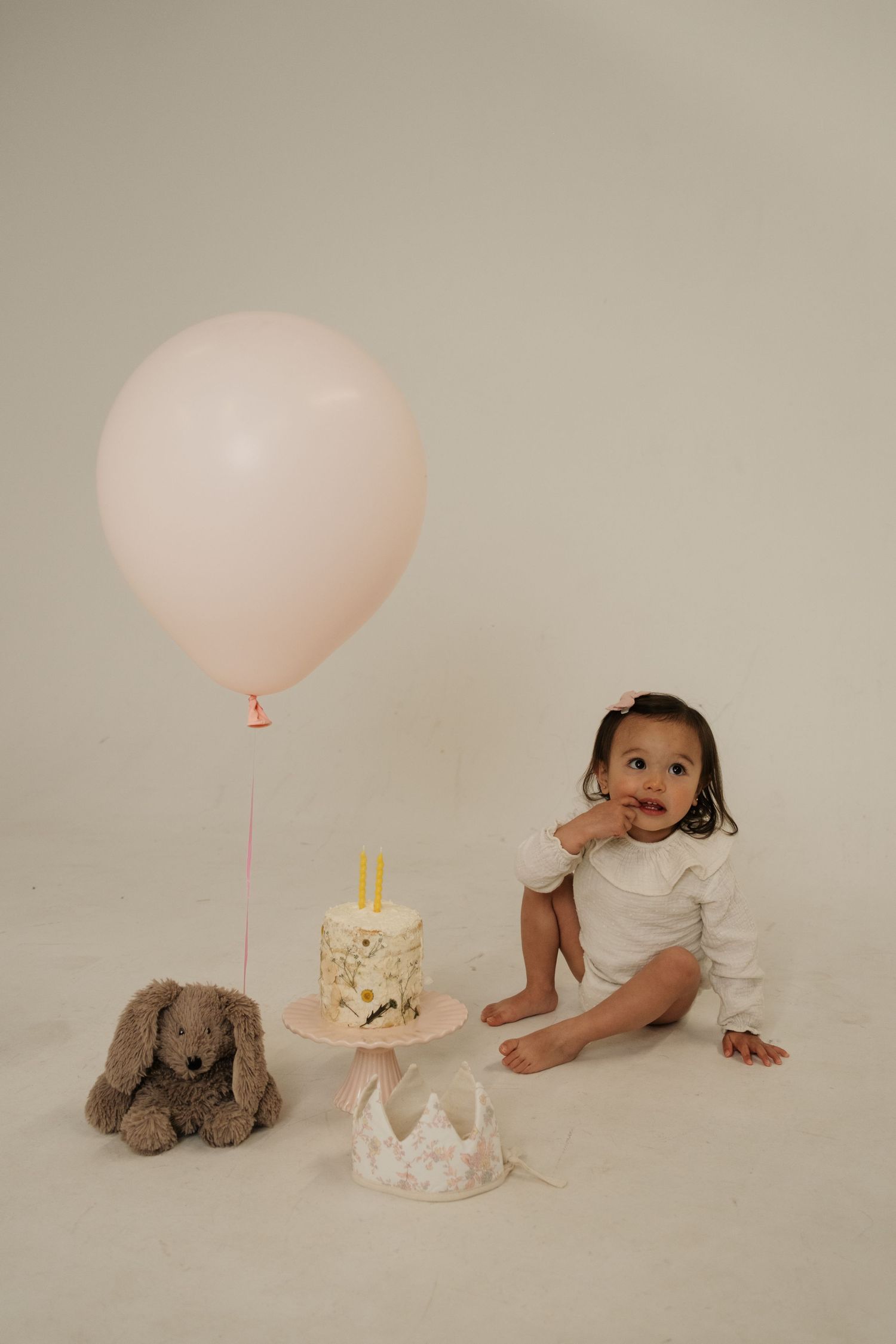 Baby girl in white onesie sits beside first birthday cake, pink balloon, and stuffed bunny on white background.
