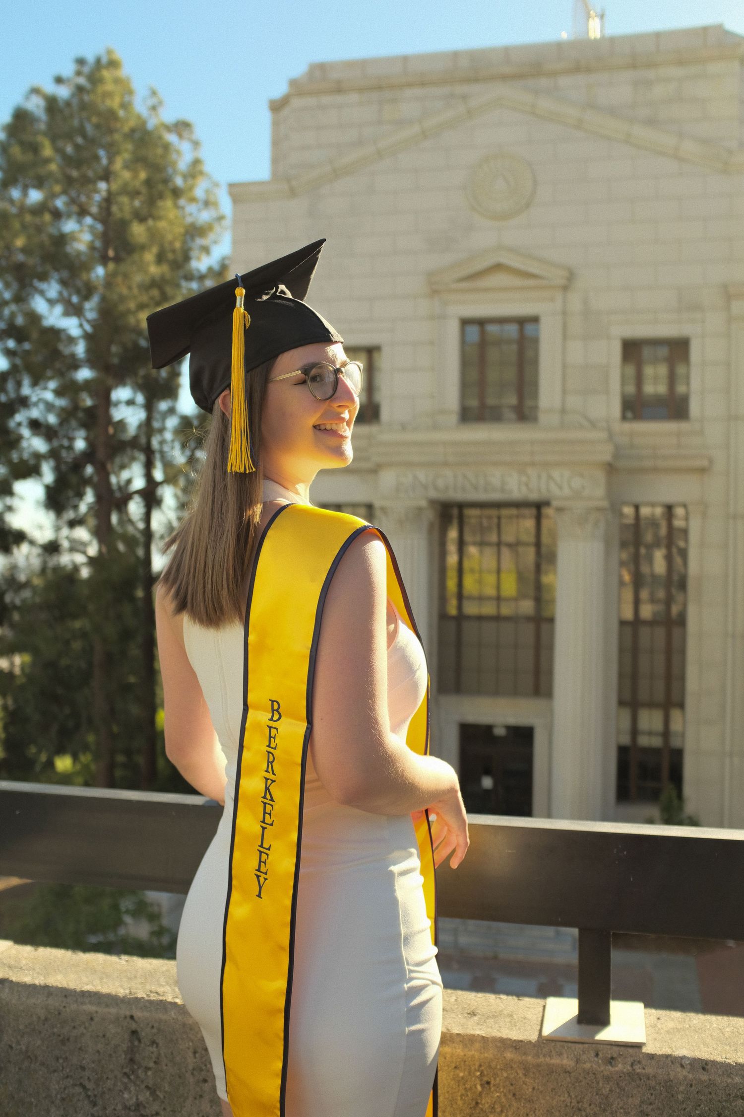 A graduate in cap and gown poses in front of a classical stone building.