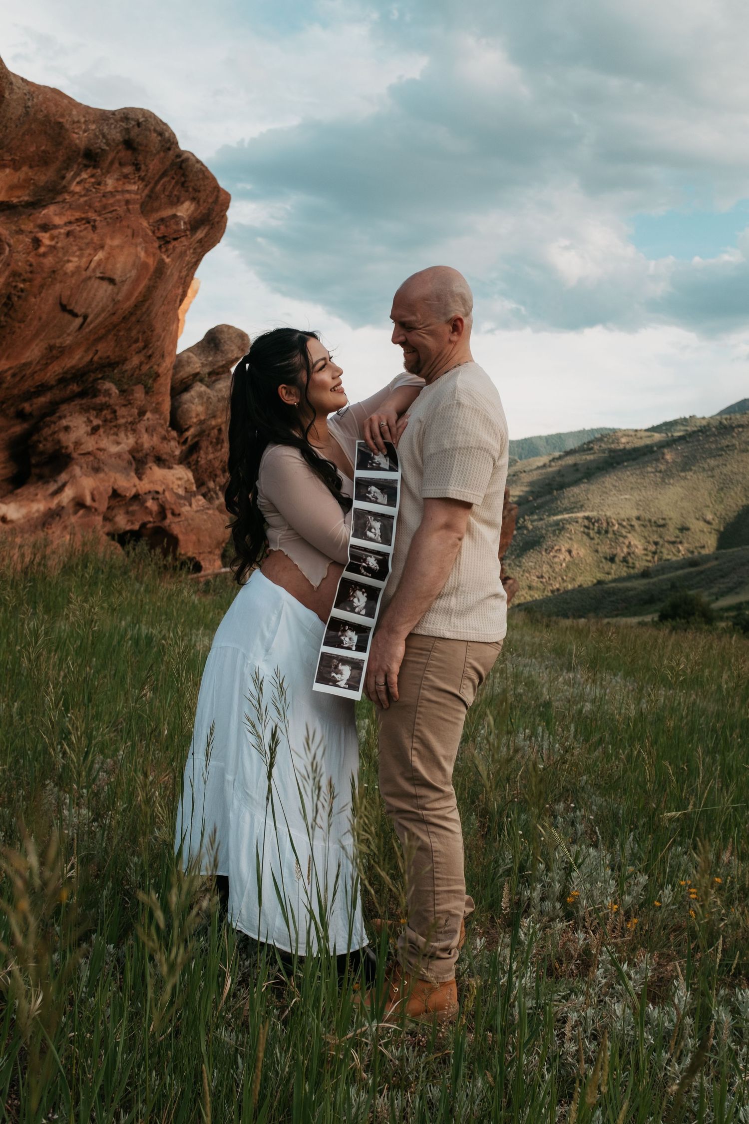 A couple stands in front of red rocks holding ultrasound photos during a pregnancy announcement photo shoot.