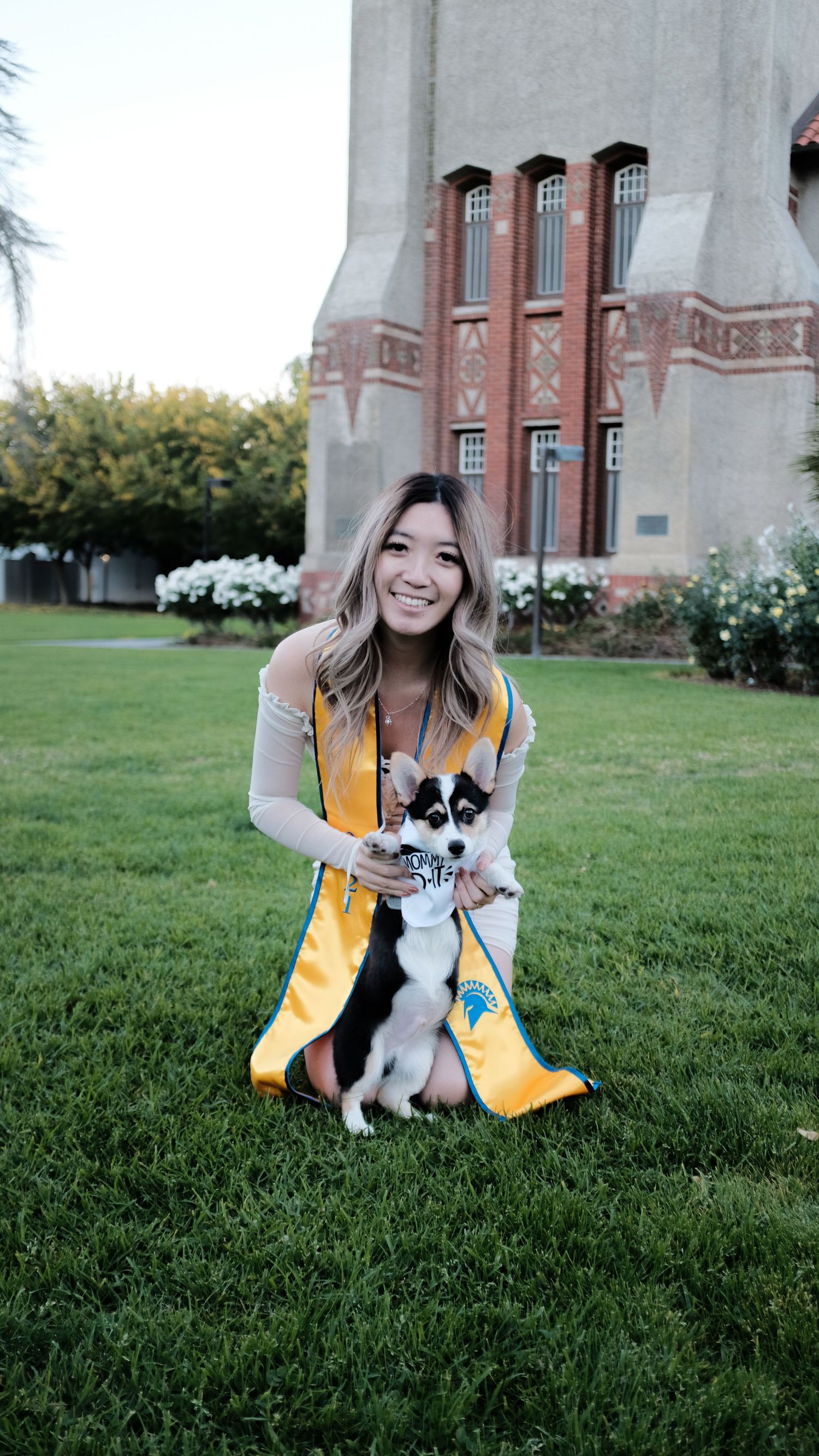 Graduate in yellow stole kneeling on grass with small black and white dog.
