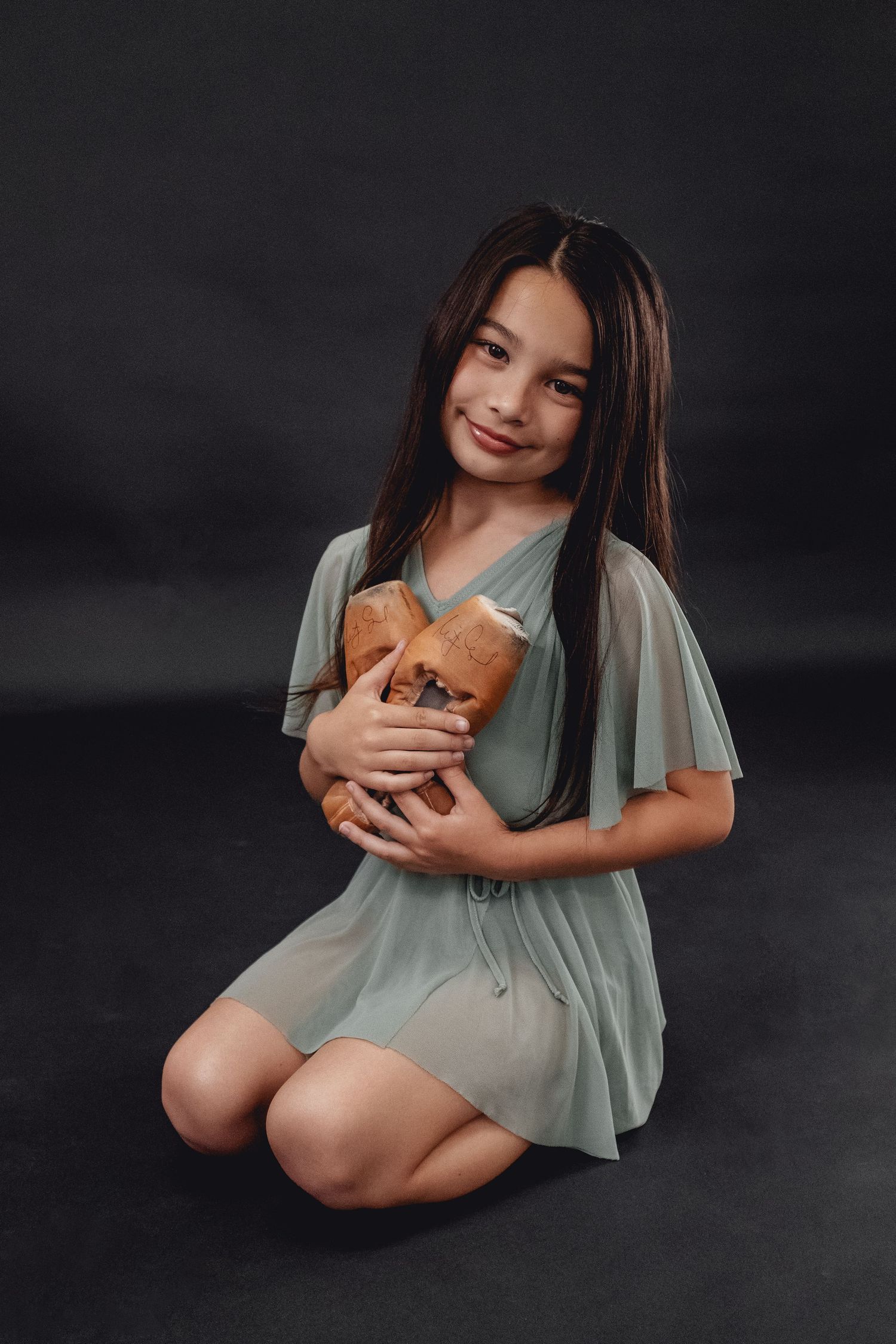 Young dancer in a flowing mint dress holds a plush toy while posing on dark background.