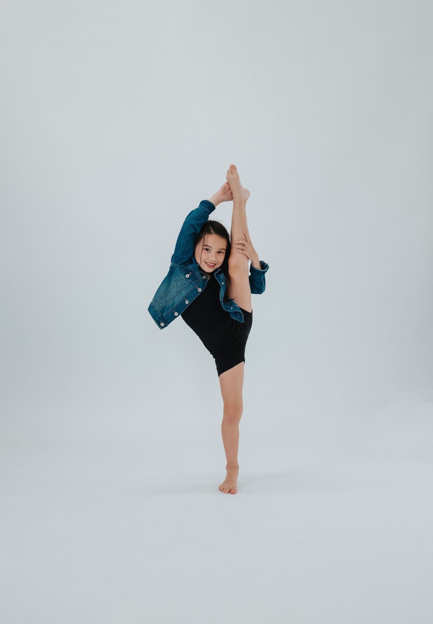 Flexible dancer performs high leg extension while wearing teal jacket in white studio setting.