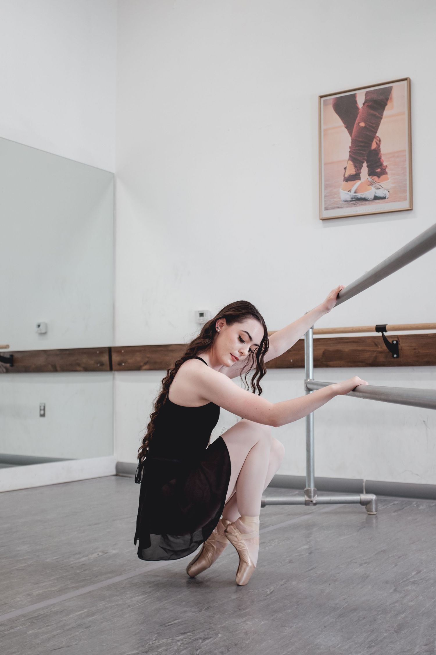 Ballet dancer in black dress practicing at barre in dance studio with mirrors and hardwood floor.