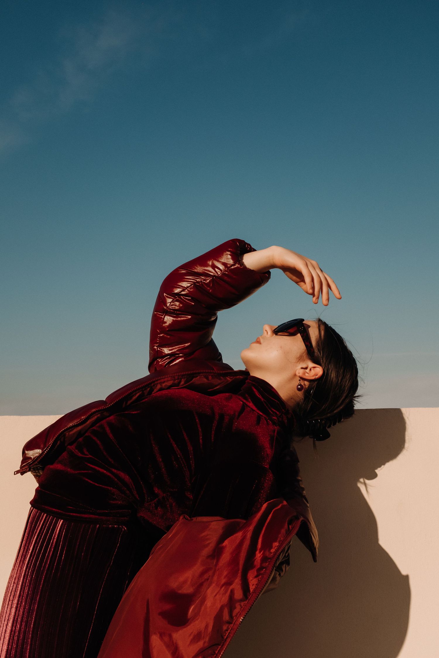 Figure in burgundy velvet outfit poses dramatically against blue sky backdrop.