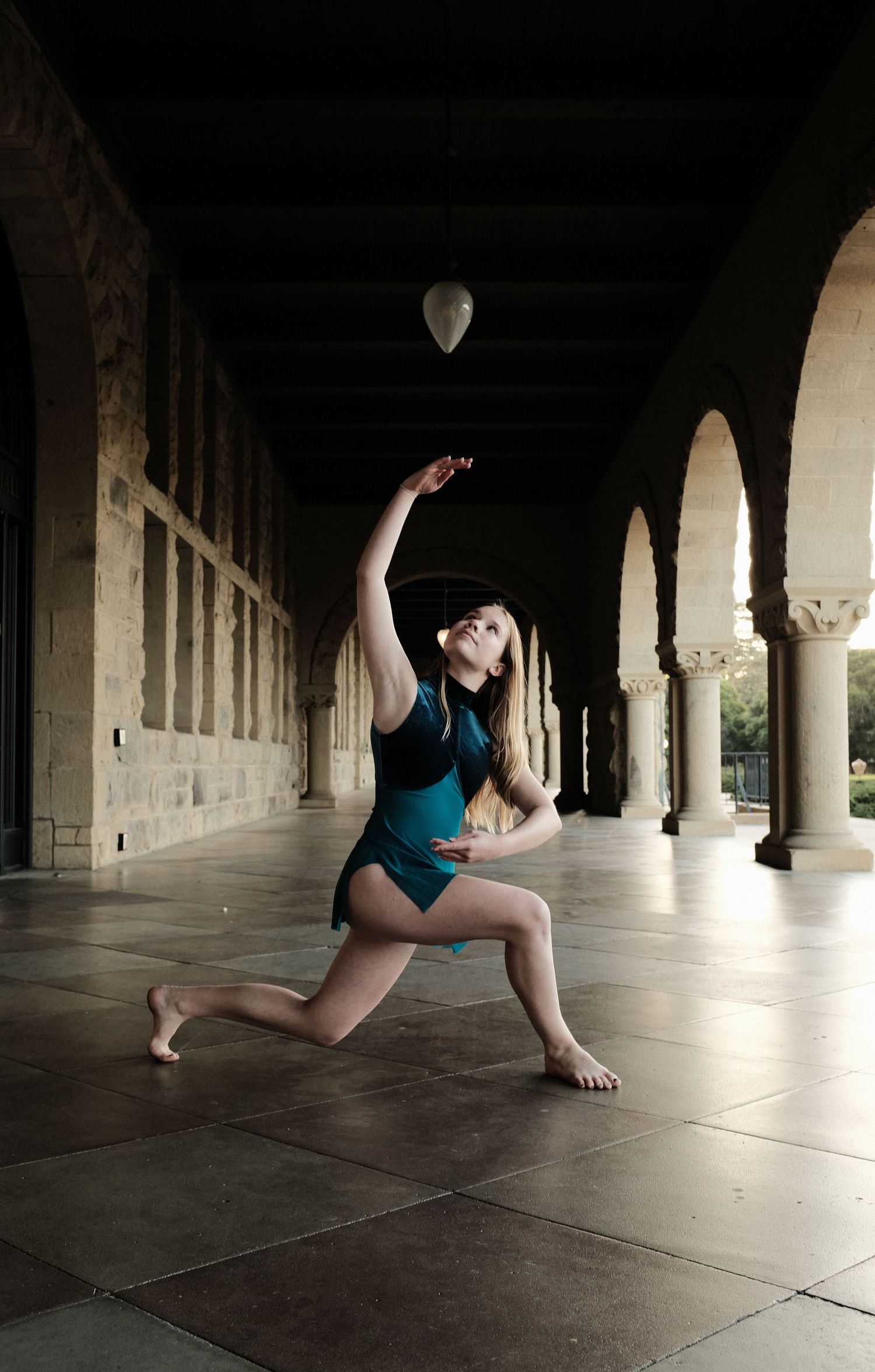 Dancer in teal leotard strikes dramatic pose in columned hallway with balloon decoration.