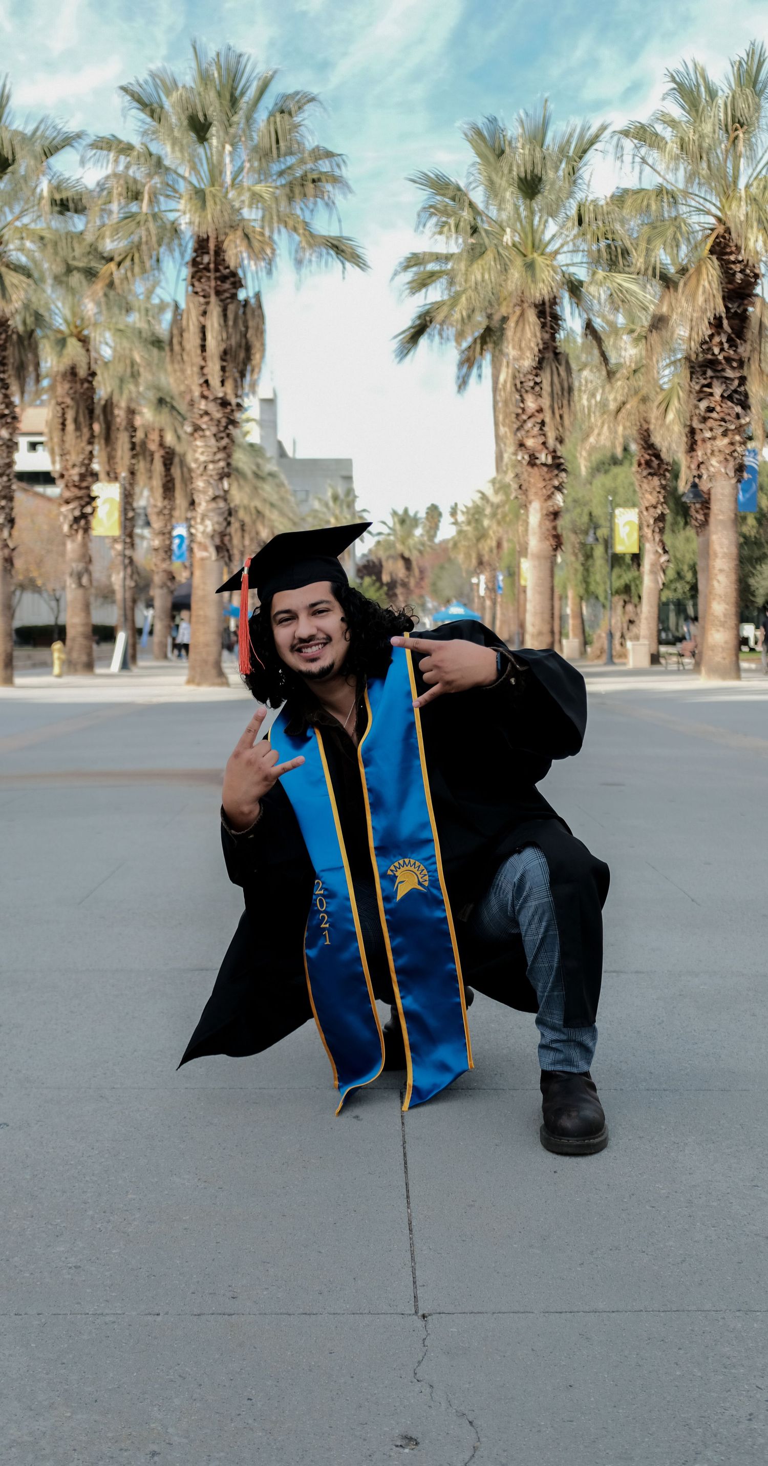 Graduate in black gown and blue stole poses playfully on concrete path lined with palm trees.