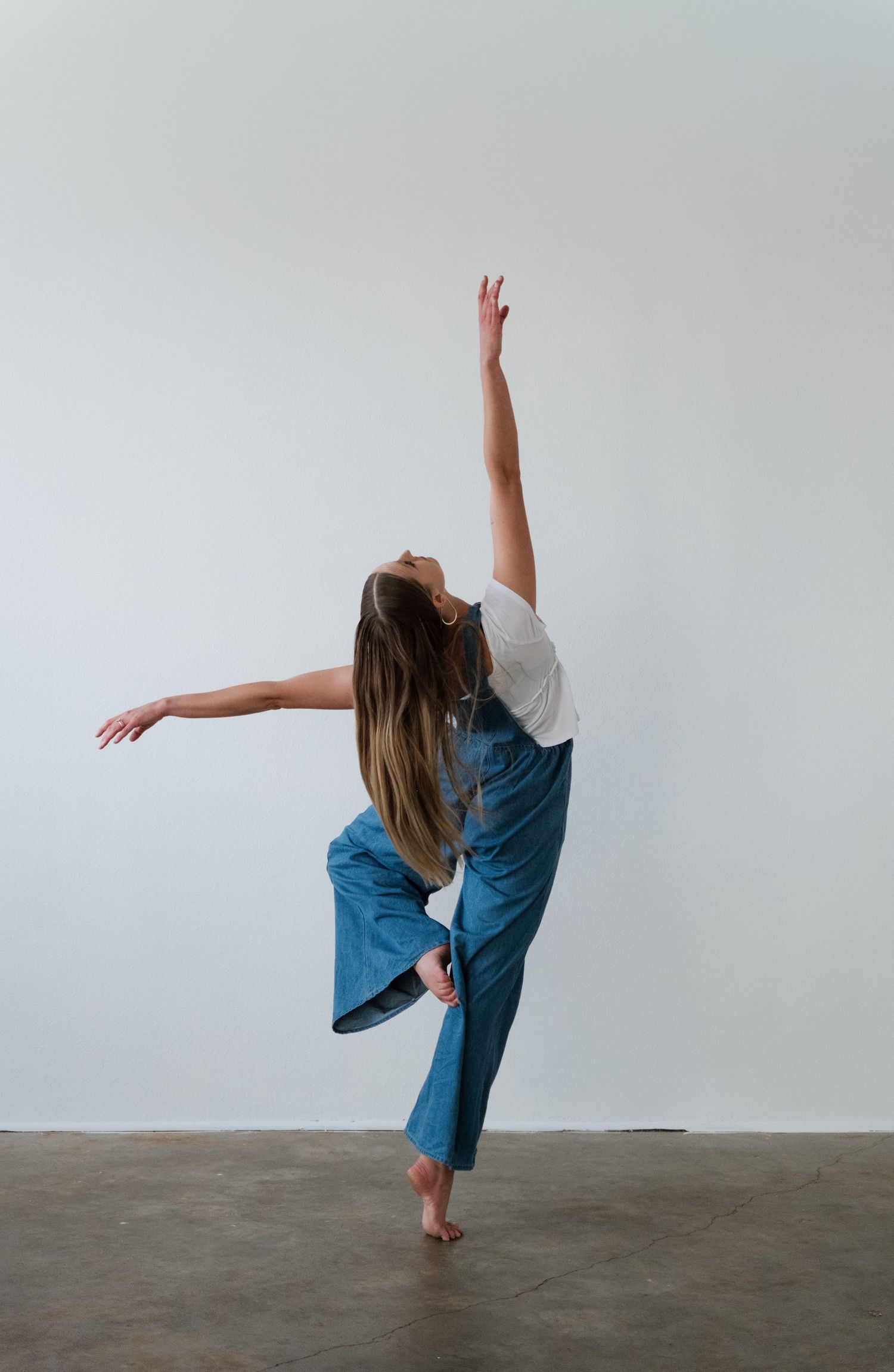 A dancer balances on one foot in blue wide-leg overalls, arms extended gracefully against a white studio wall.