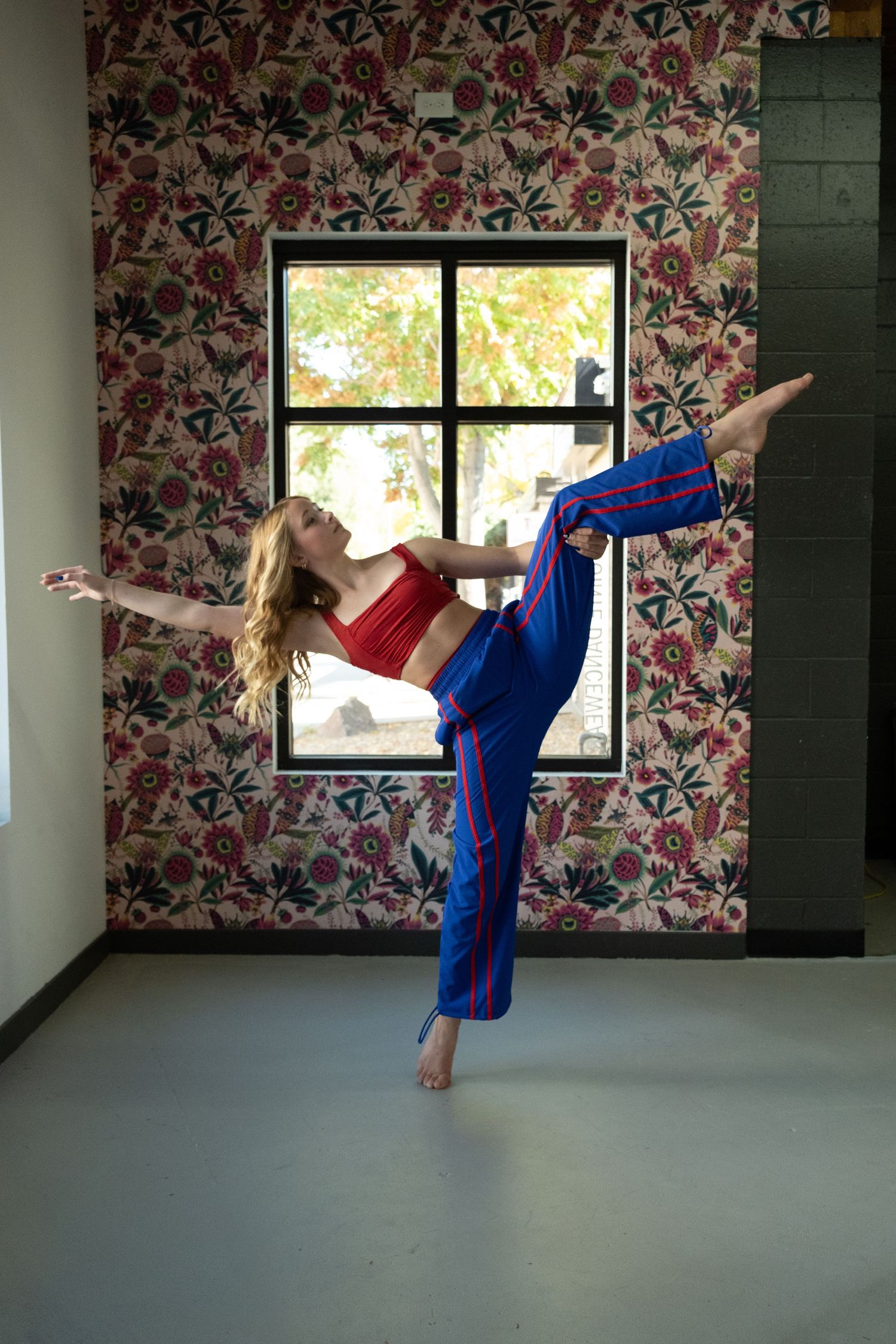 A dancer balances on one foot with leg raised high in a floral-wallpapered studio, wearing red top and blue striped pants.