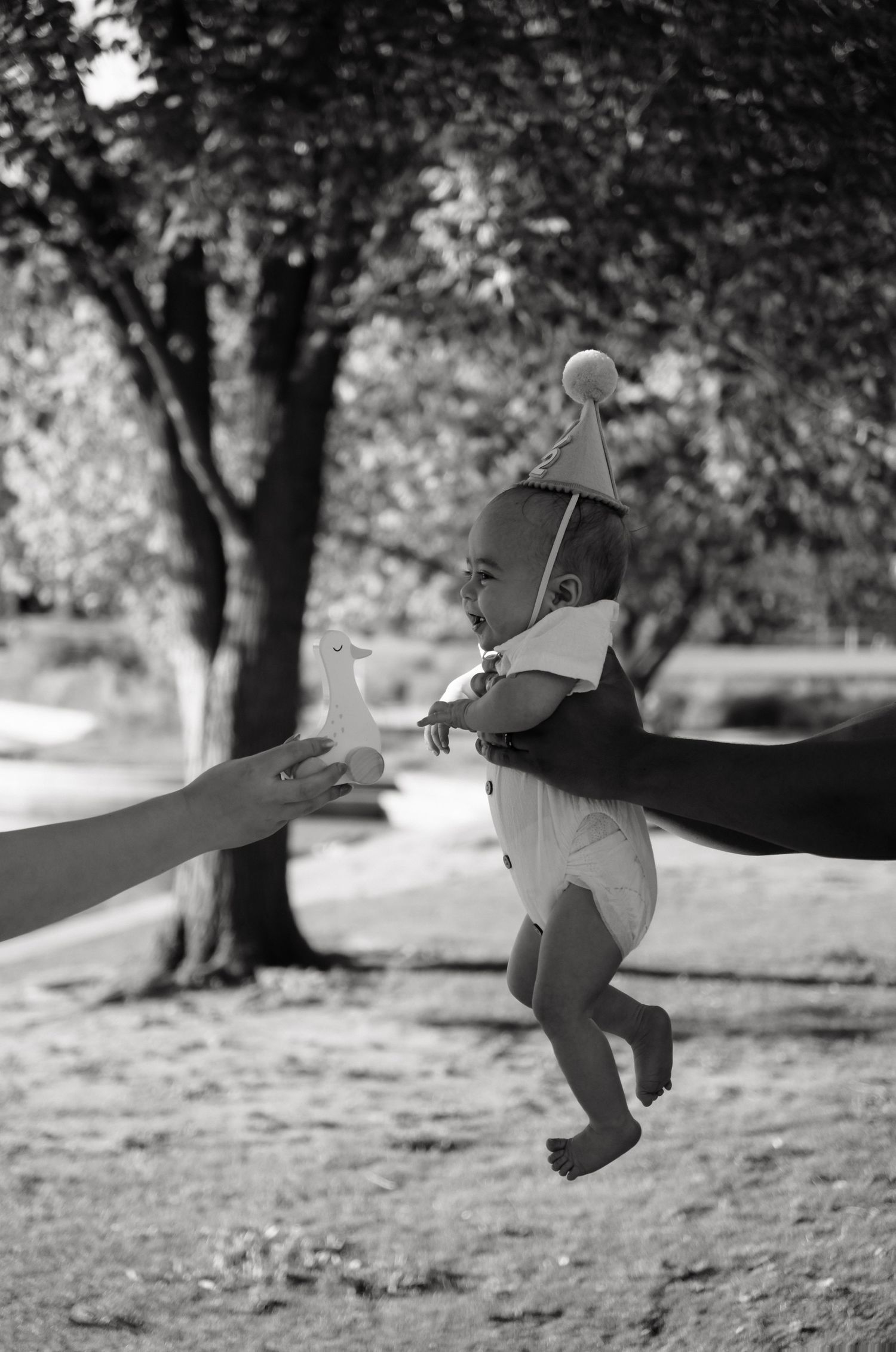 Black and white photo of a baby in a party hat being held outdoors near trees, reaching toward an outstretched hand.