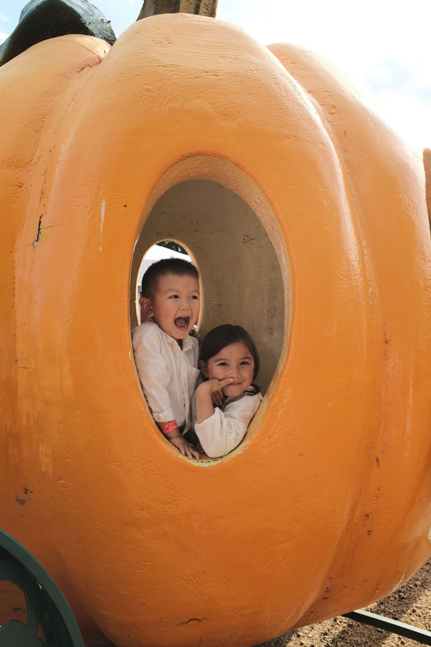 Two young children laughing and posing inside a large decorative orange pumpkin cutout at a fall festival.