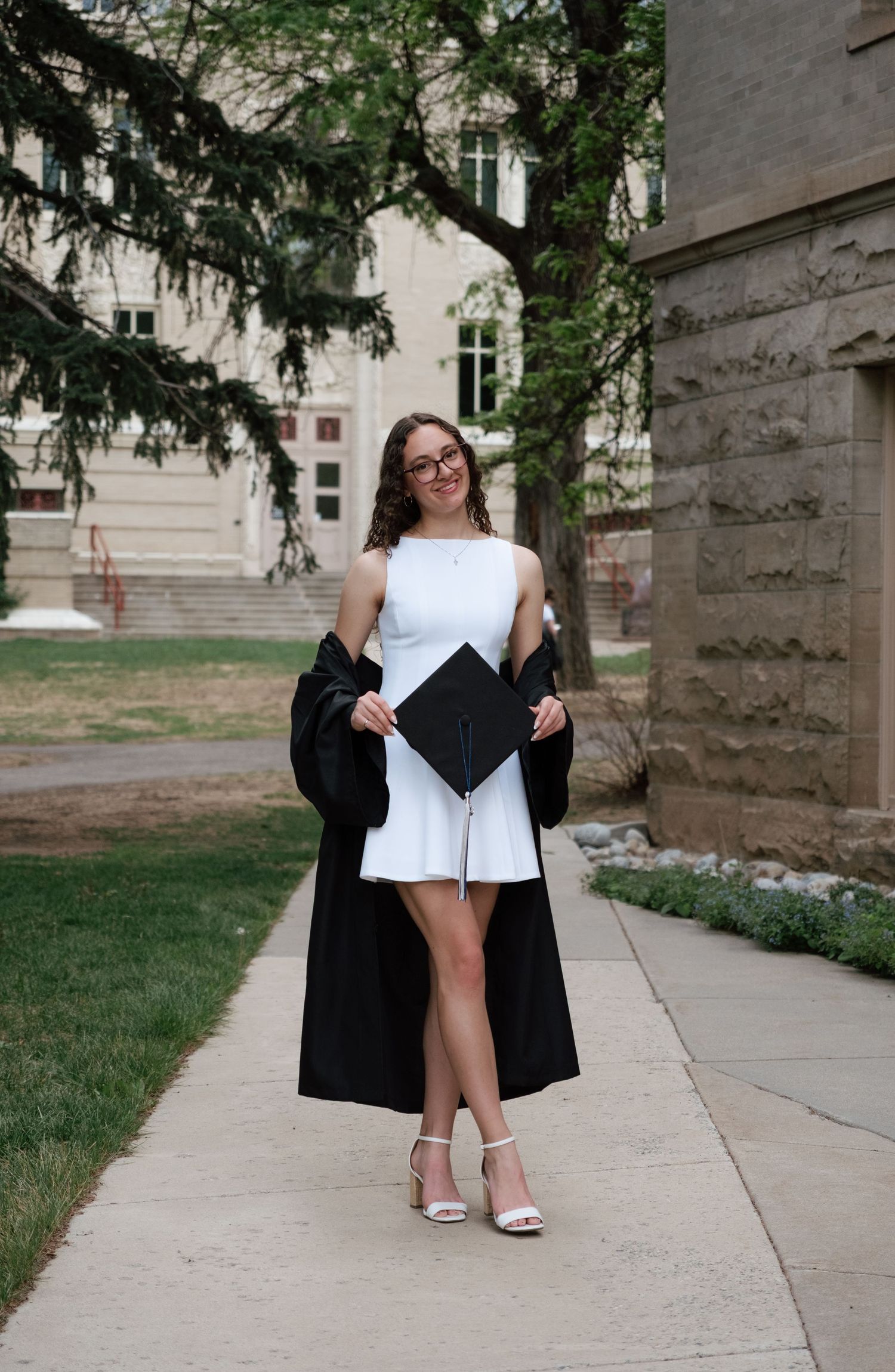 A smiling graduate in a white dress holds her cap and black gown on a campus walkway.