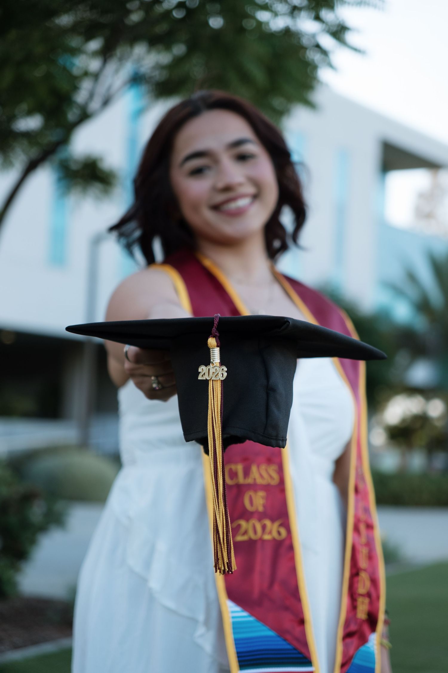 Smiling graduate holds black cap with gold tassel and Class of 2026 text, wearing white dress and maroon graduation stole.