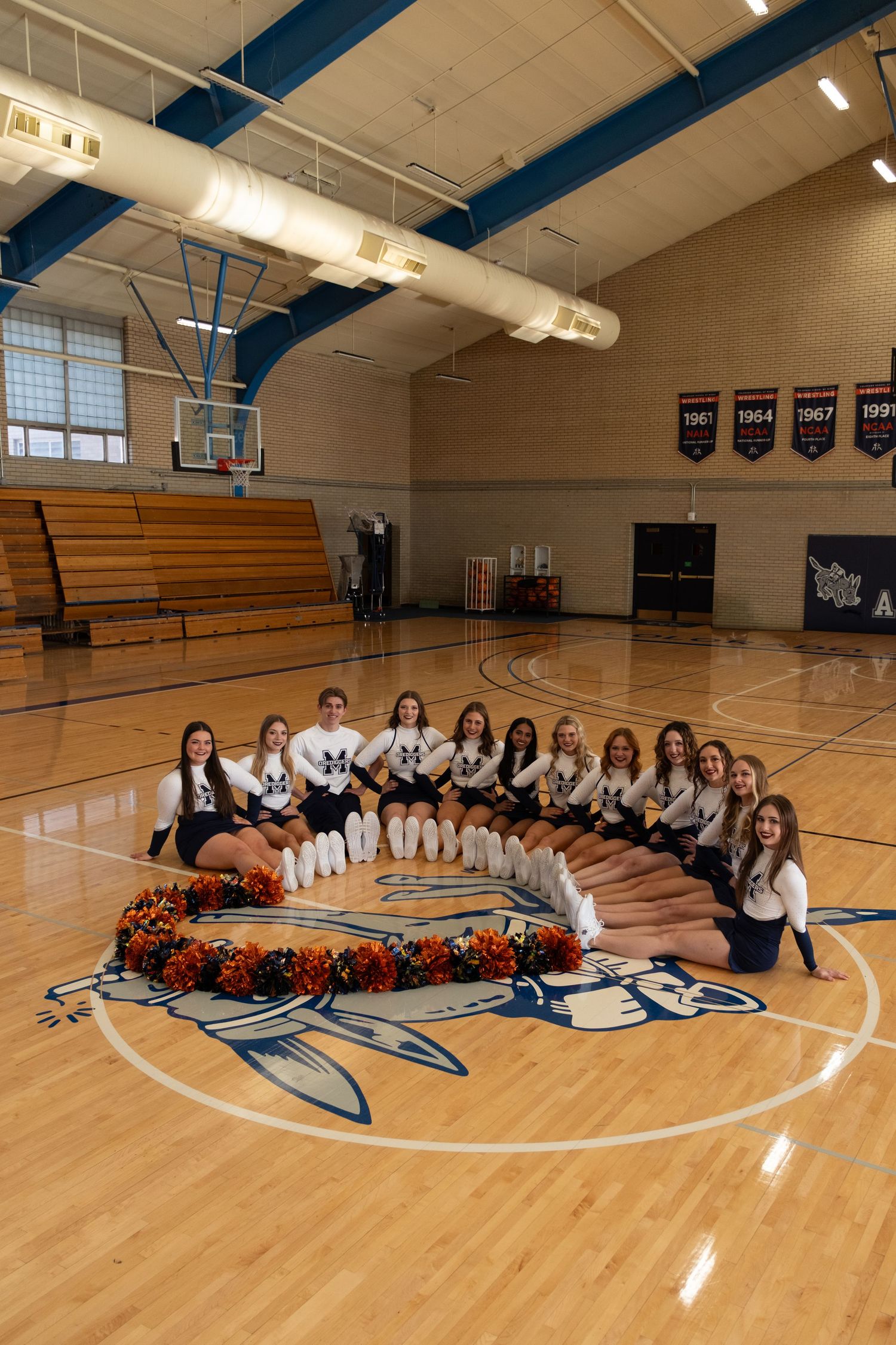 High school cheerleaders pose in a circle formation on a gymnasium floor around orange pom-poms near center court logo.