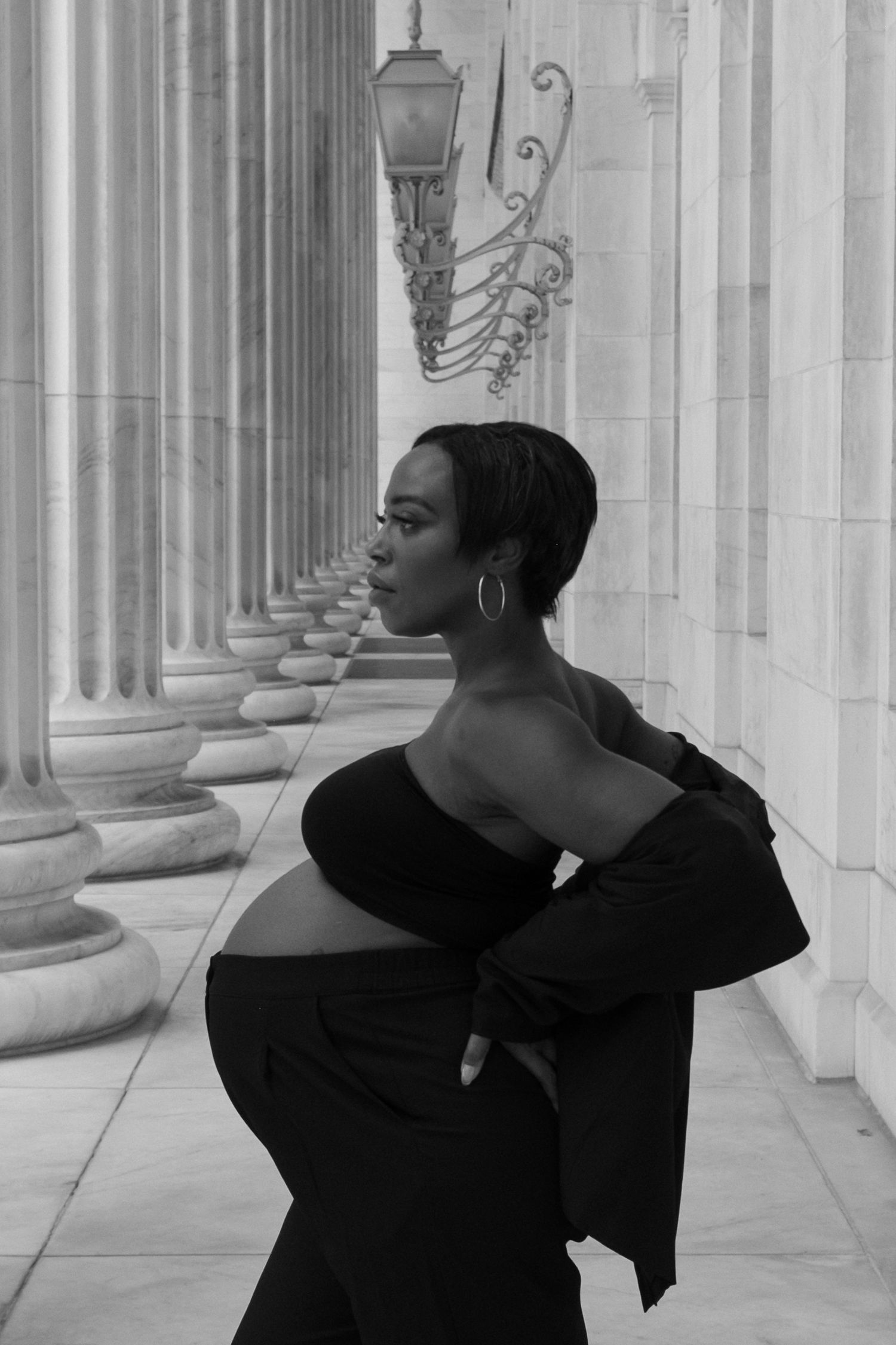 Black and white photo of a pregnant woman in an off-shoulder black dress posing elegantly among classical stone columns.