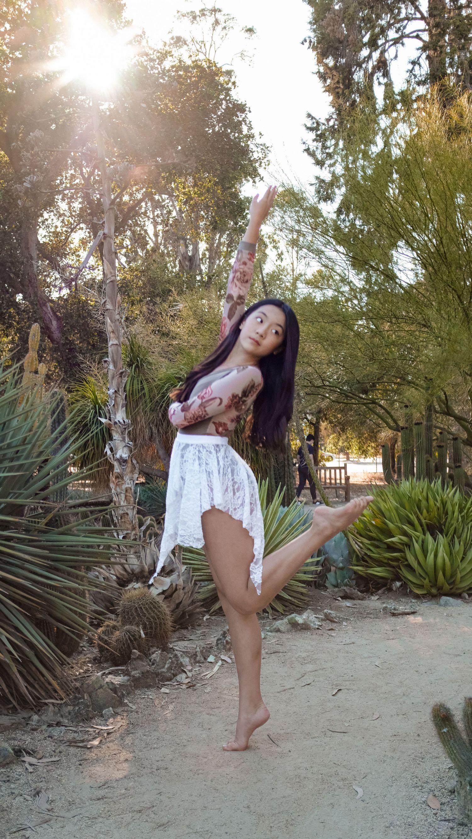Dancer strikes elegant pose in sunlit garden path surrounded by desert plants.