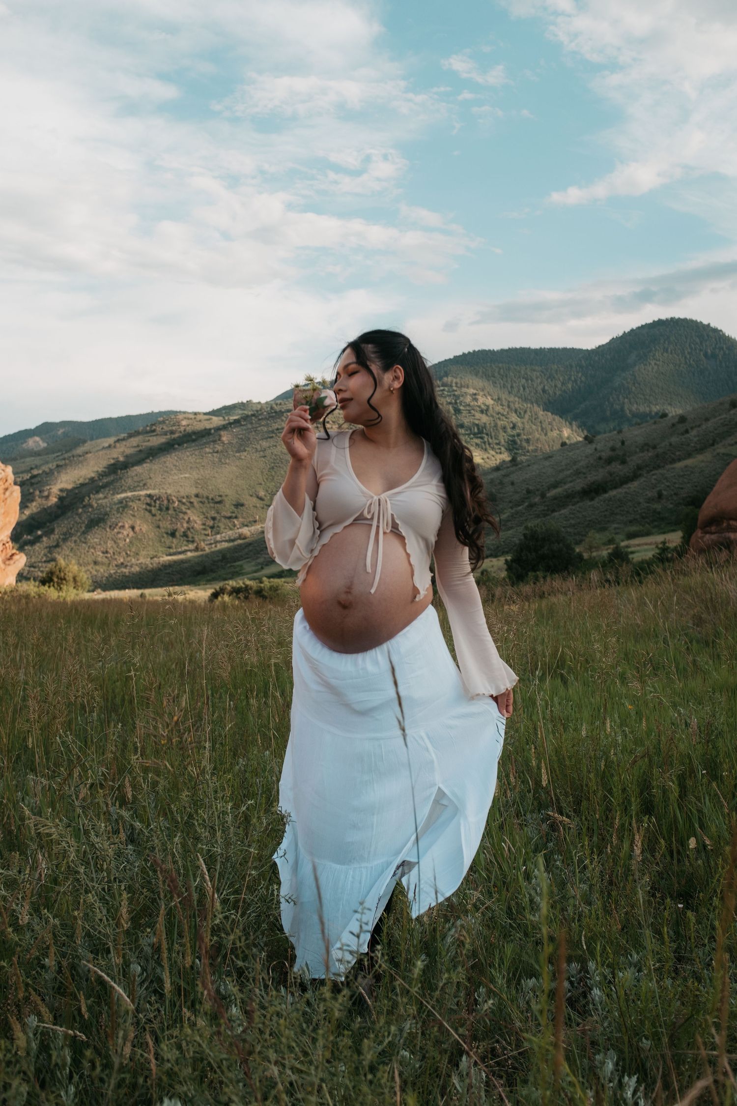 Pregnant woman in white skirt and sheer top smelling wildflowers in a green meadow with mountain backdrop.