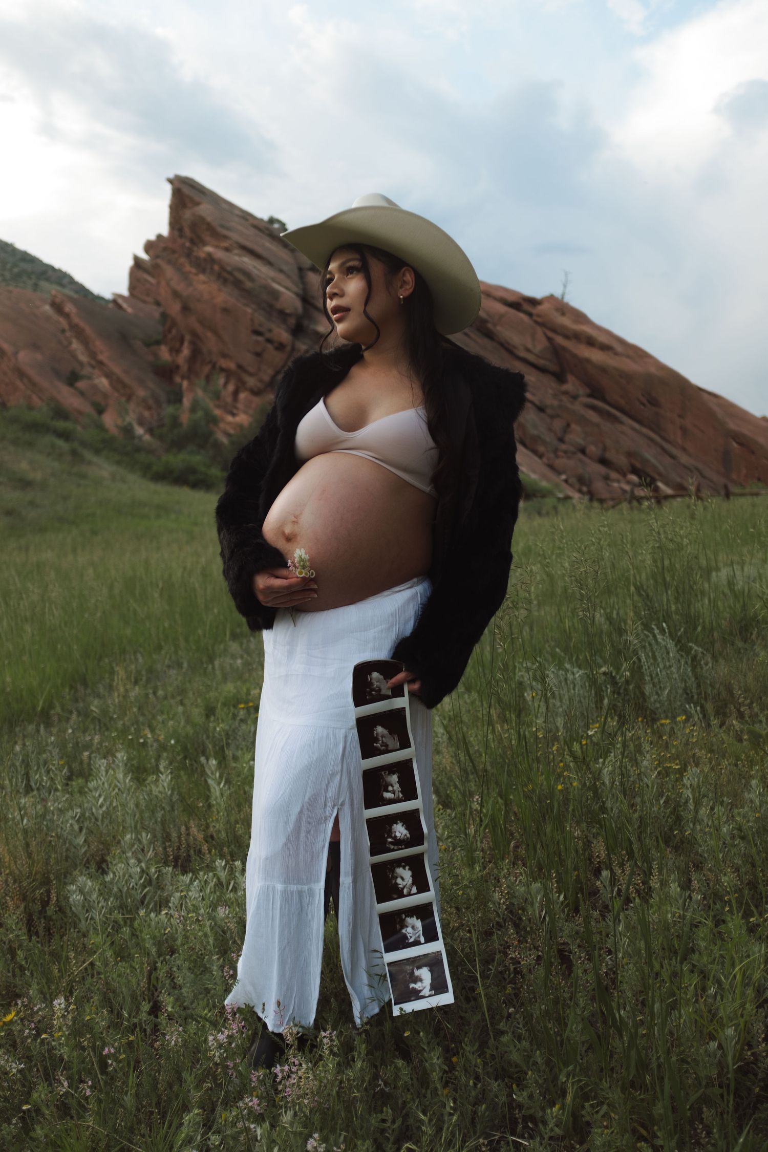 Pregnant woman in fur coat and cowboy hat holds ultrasound strip in field near red rocks.