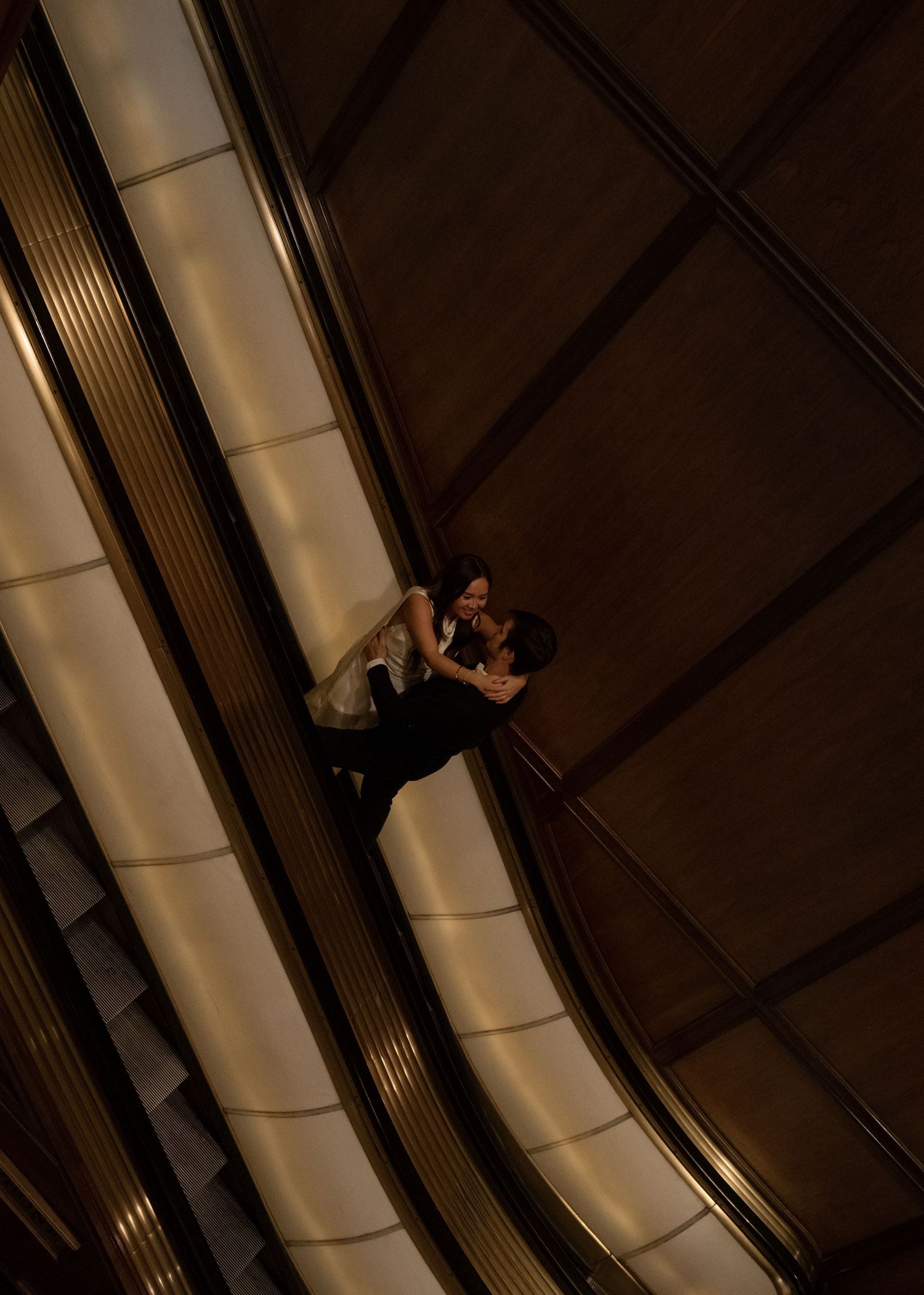Couple sharing a romantic embrace on a grand escalator in a dimly lit luxury building atrium.