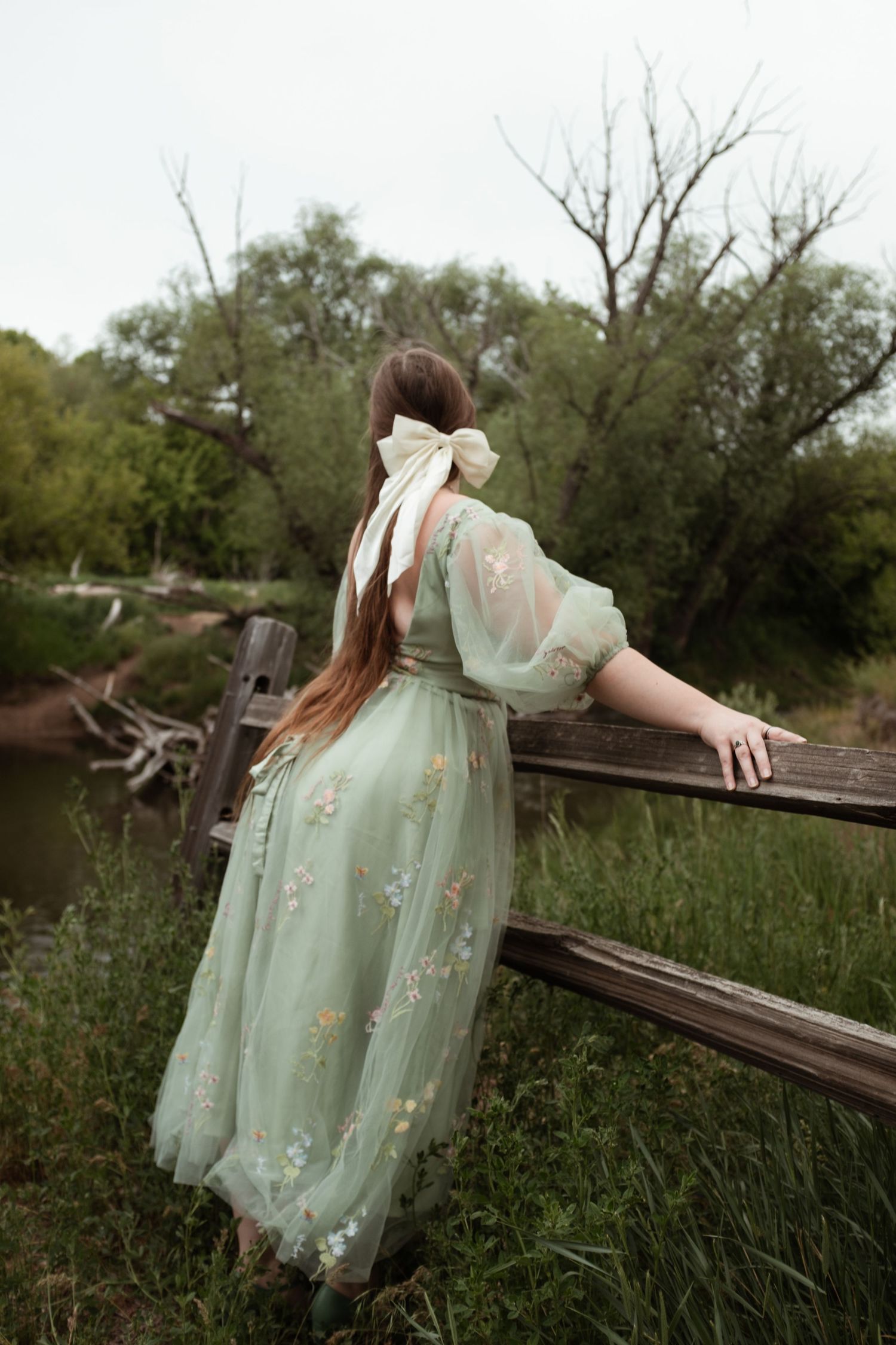 Person in ethereal mint gown stands dreamily at wooden fence overlooking natural landscape.