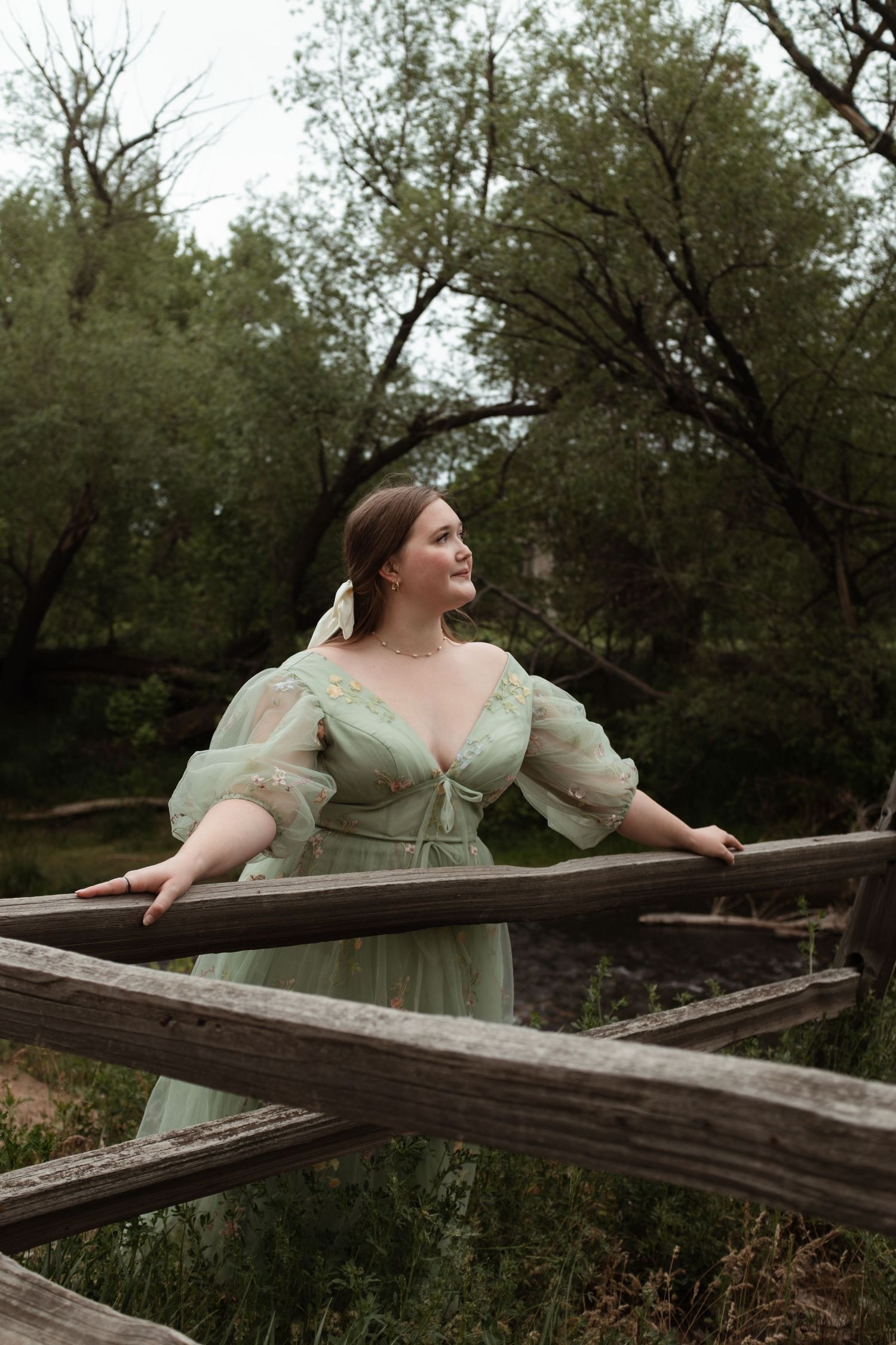 A person in a light green vintage-style dress poses dramatically along a wooden fence in a wooded setting.