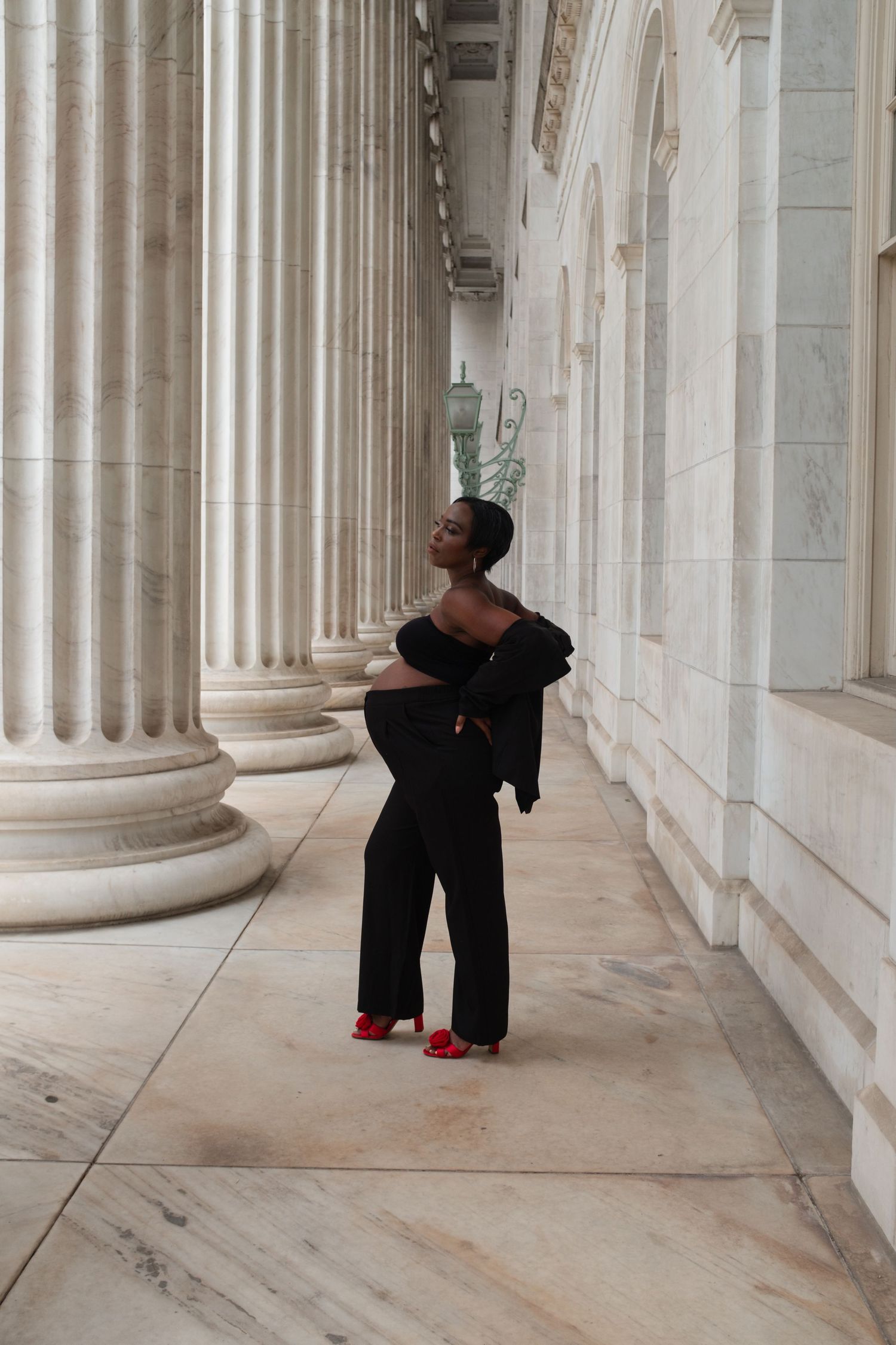 Woman in black off-shoulder jumpsuit and red heels posing elegantly among grand marble columns.