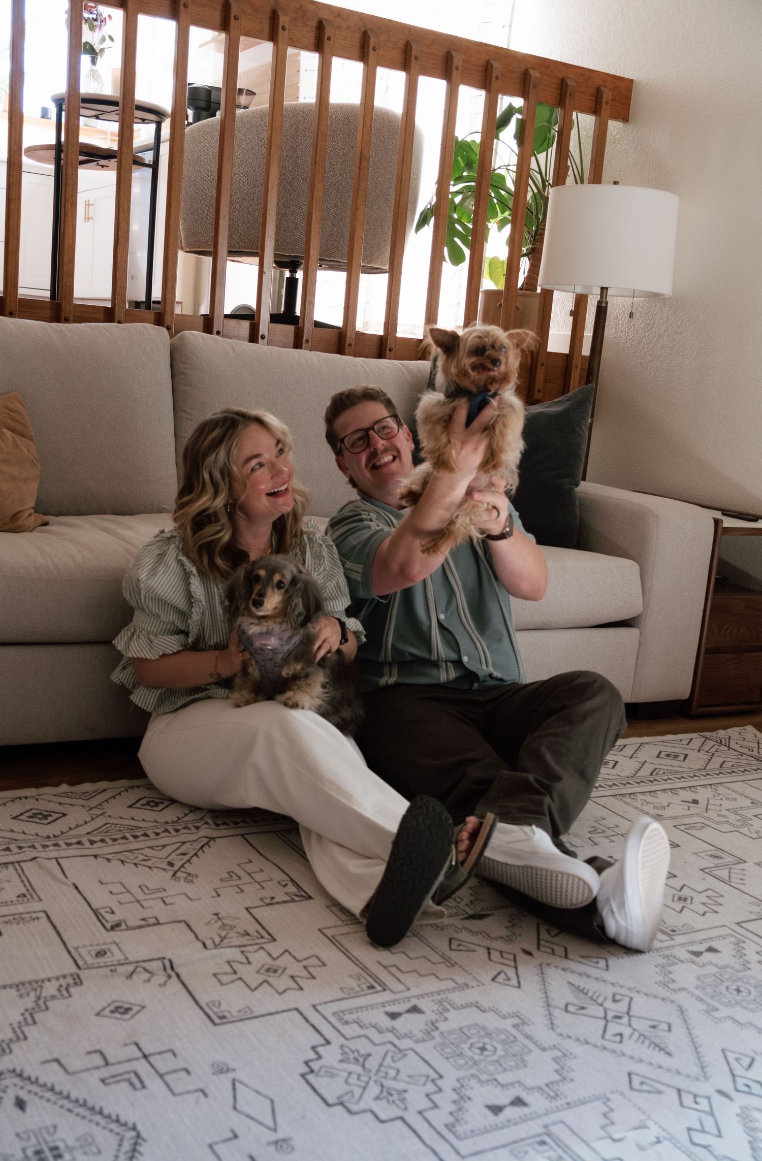 A smiling couple sits on a patterned rug in a cozy living room, holding up one small dog while another dog rests beside them.