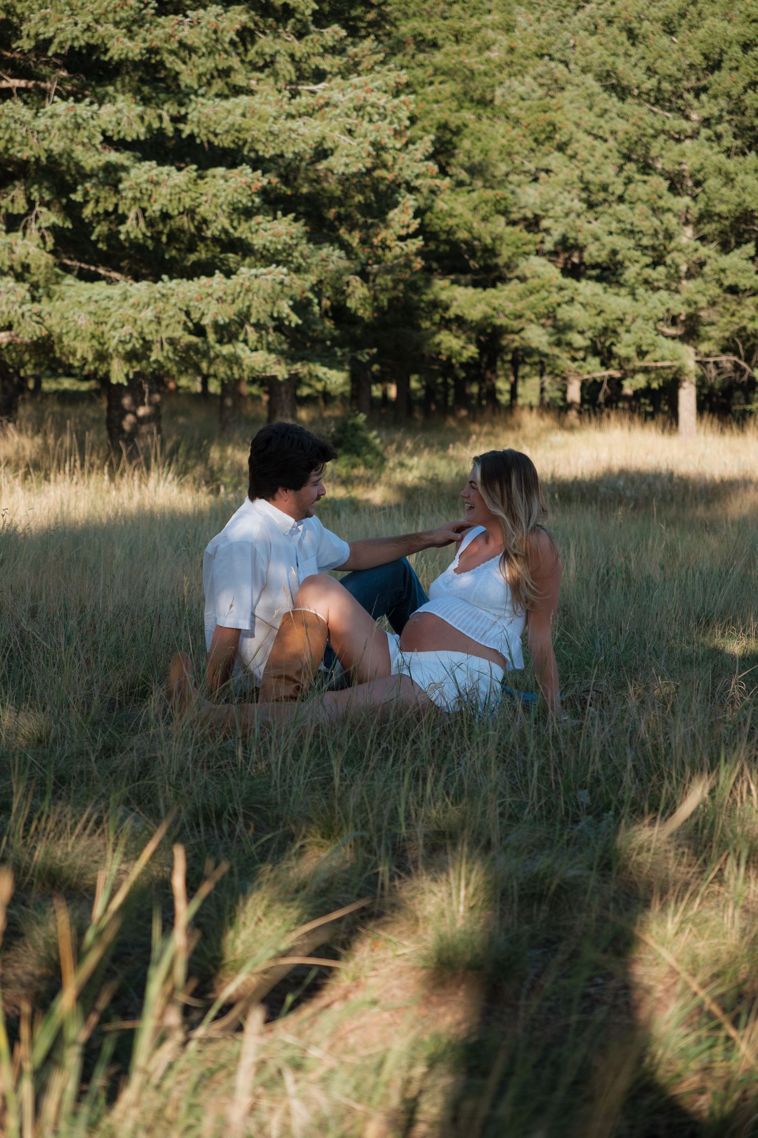 Expectant couple in white outfits sit together in a sunlit grassy meadow with tall evergreen trees behind them.