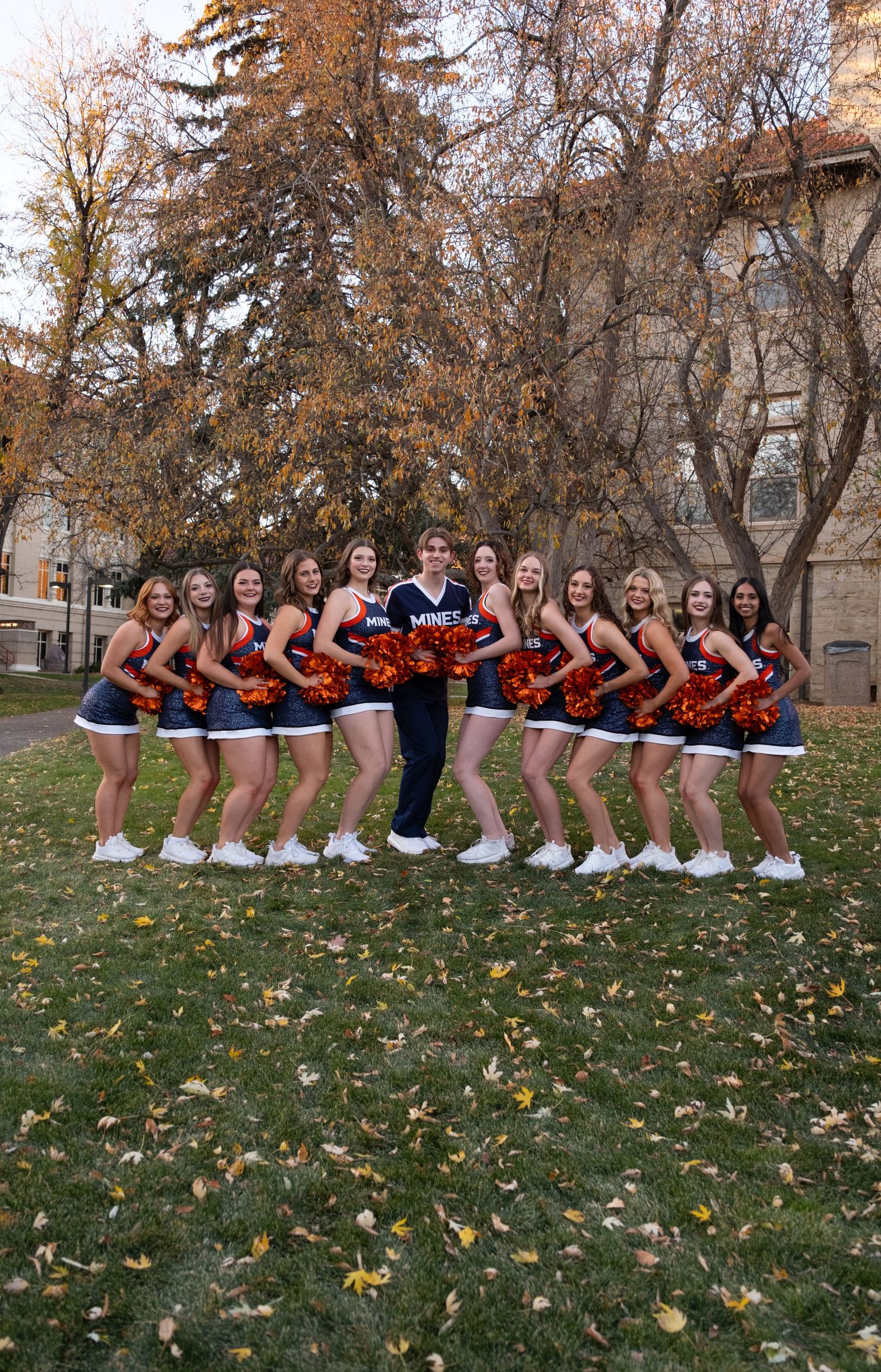 College cheerleading squad posing outdoors in blue and orange uniforms with pom-poms on a fall campus lawn.