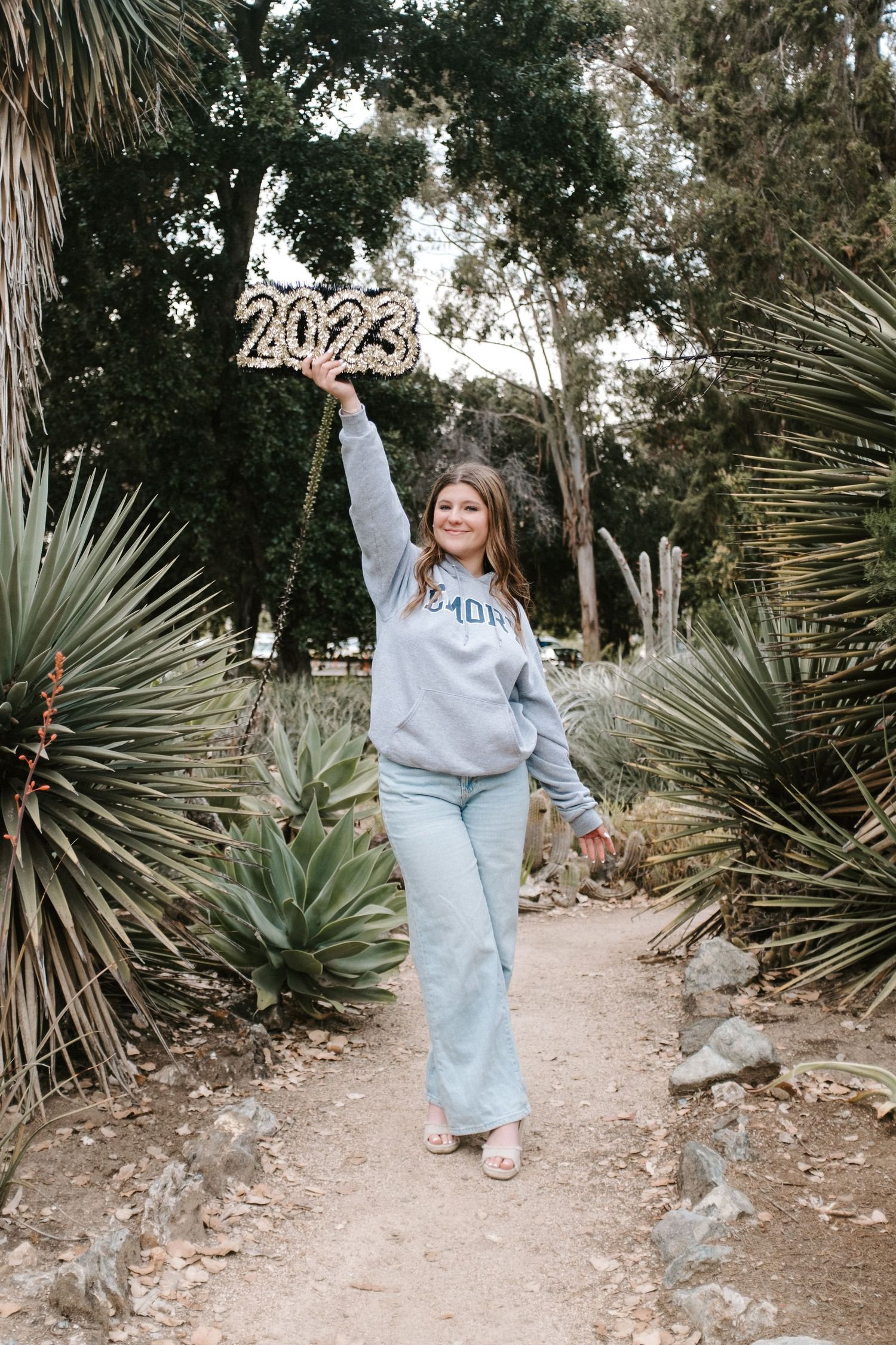 Graduate in light gray sweatsuit poses cheerfully in desert garden with palm trees.