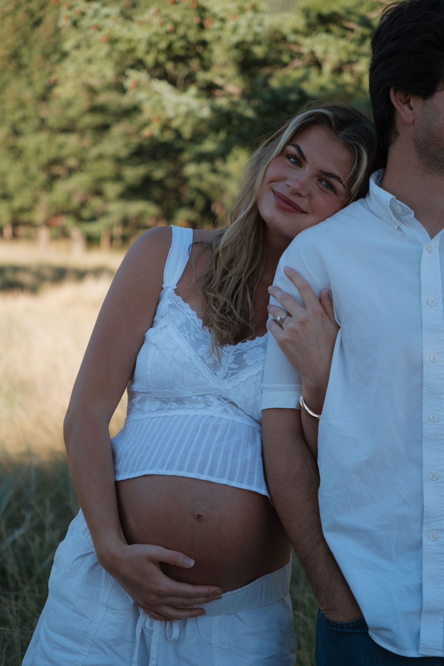 Pregnant woman in a white lace top resting her head on a man's shoulder, hand on her belly, in a sunlit grassy field with trees.