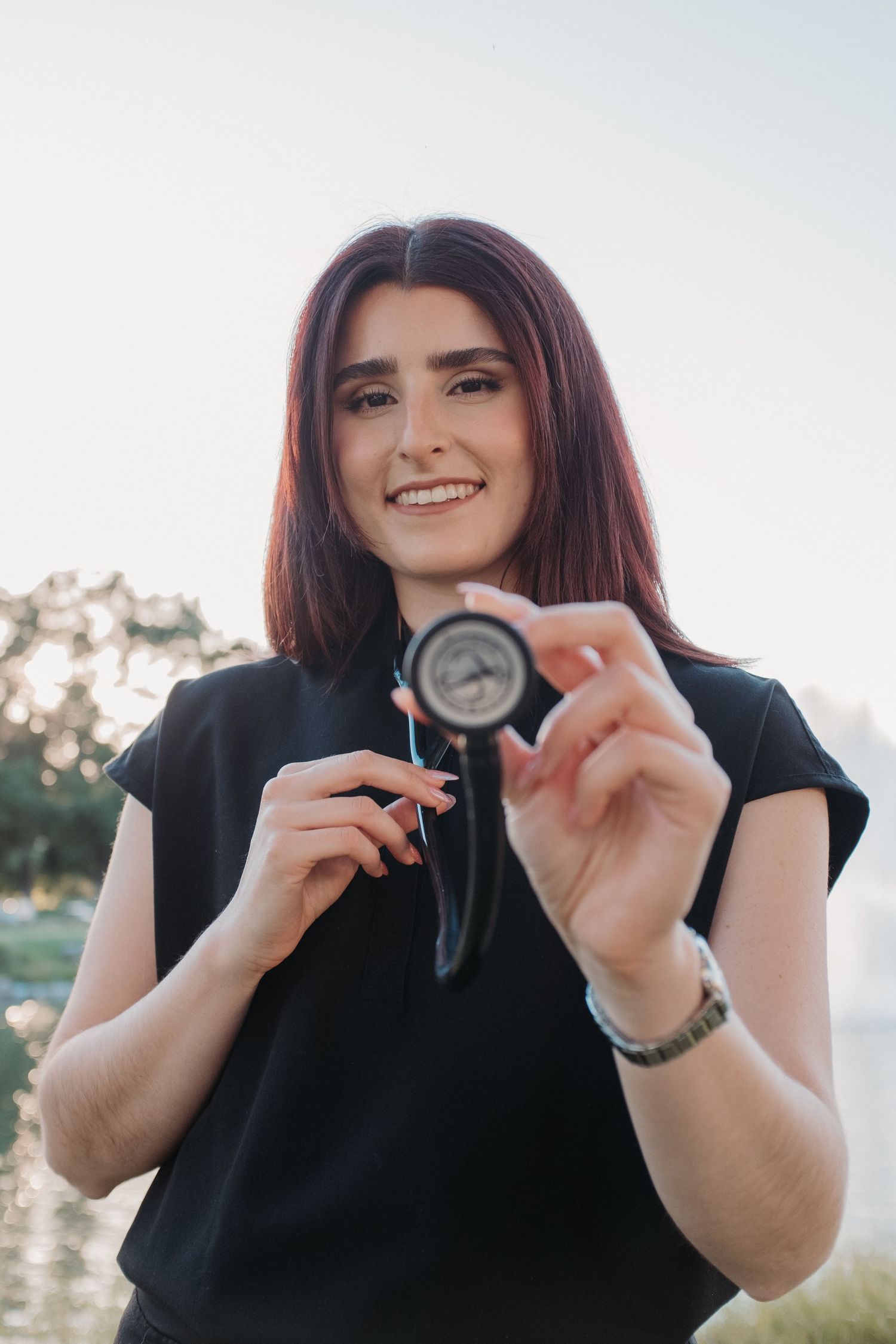 Healthcare graduate in black attire holding a stethoscope outdoors.