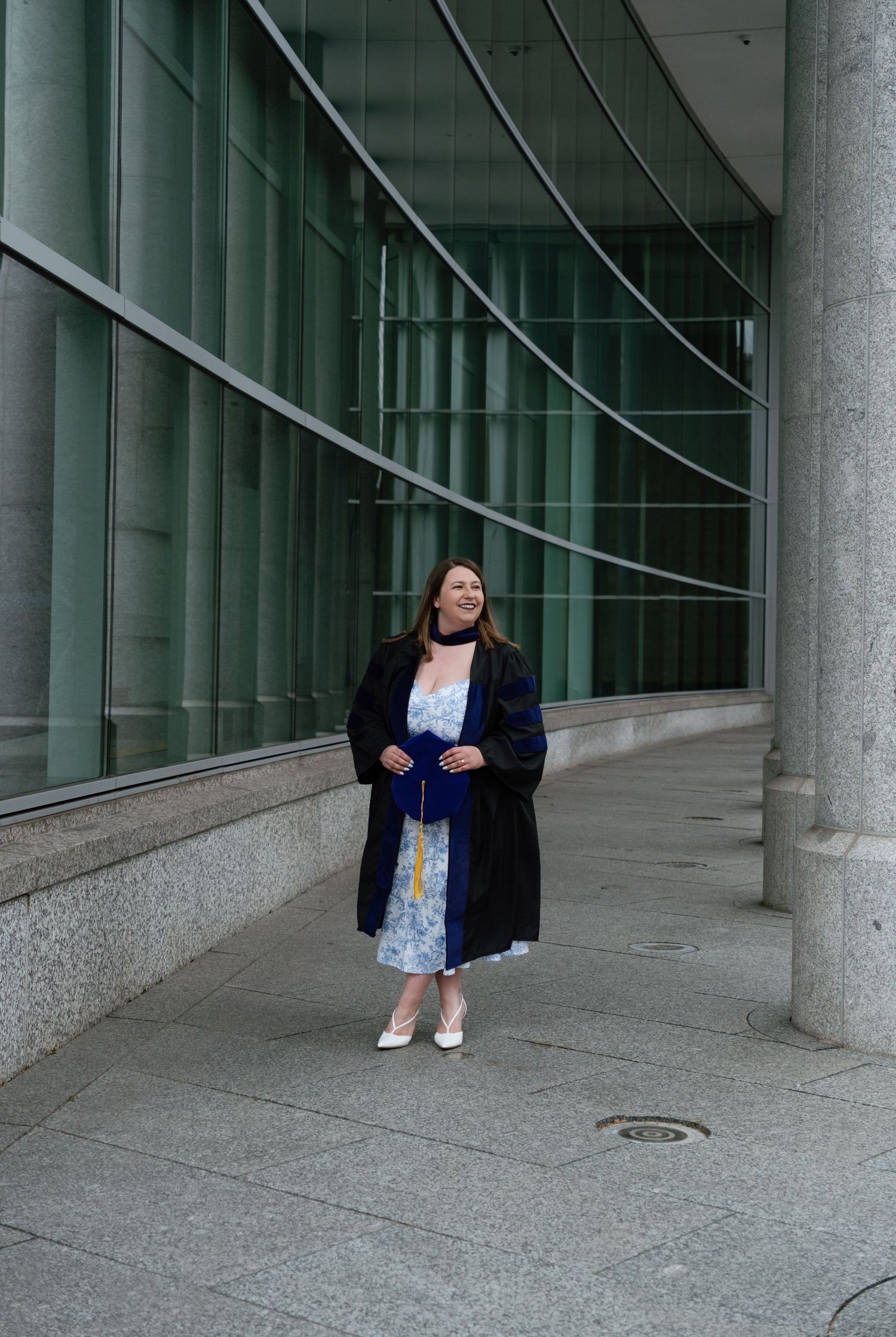 A woman in graduation gown and floral dress stands smiling outside a modern glass building.