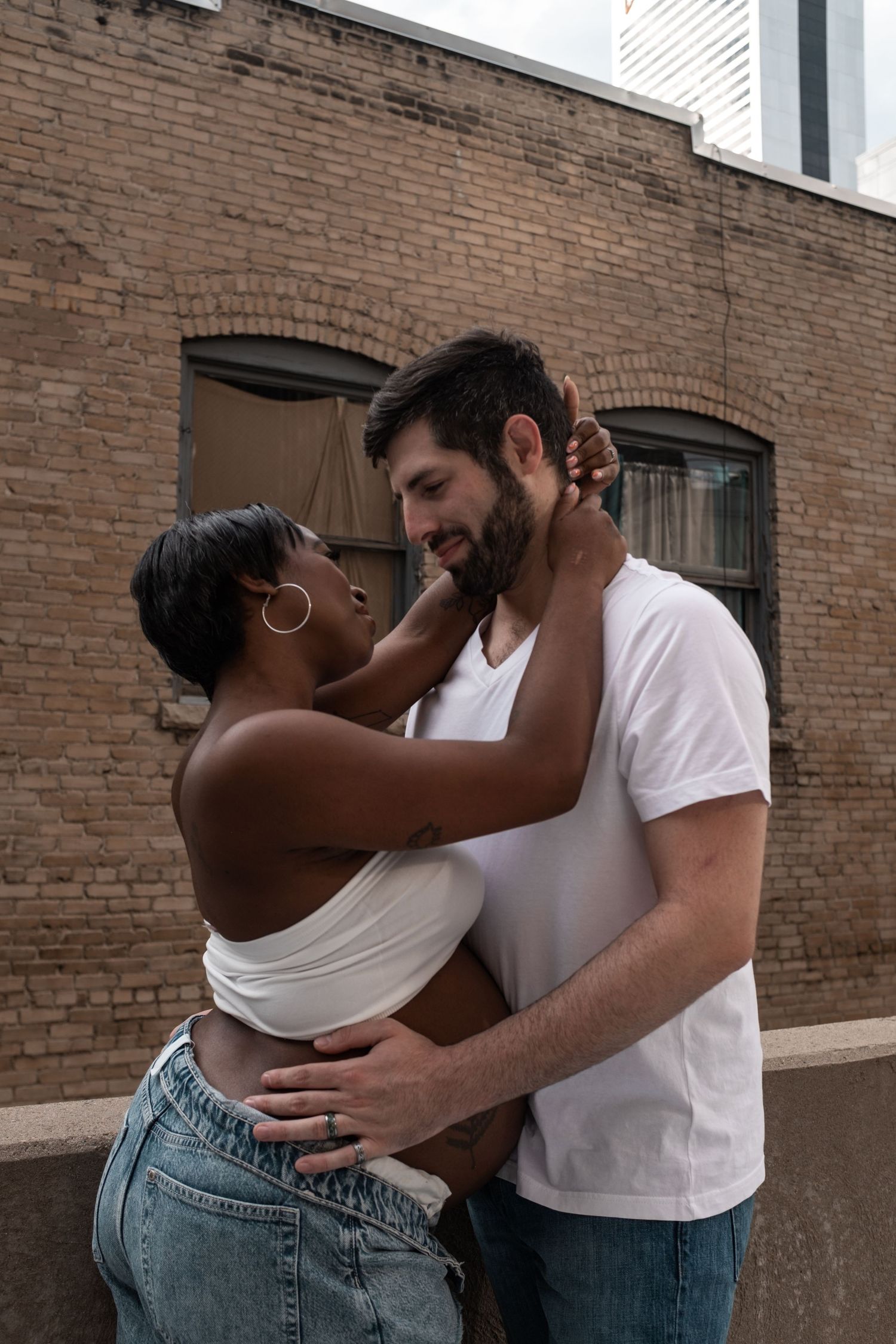 Interracial couple embracing romantically outdoors against a brick building, woman touching man's face tenderly.