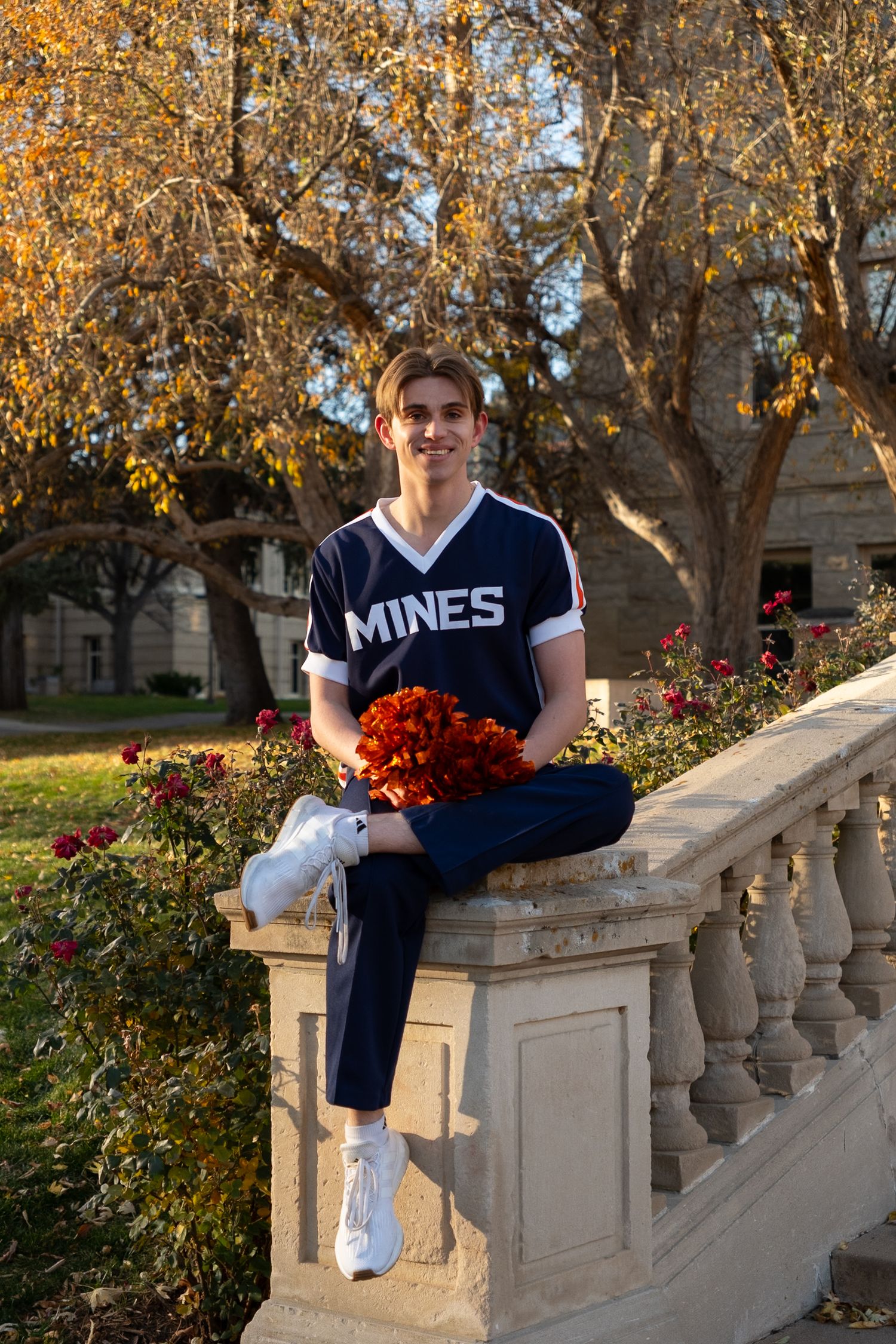 Young cheerleader in Mines jersey sits on stone railing holding red pom-poms surrounded by autumn trees.
