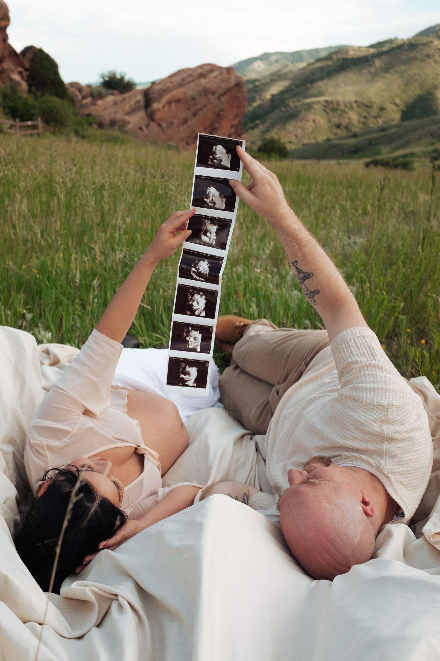 A couple lying on a blanket examines ultrasound photos against a backdrop of mountains.