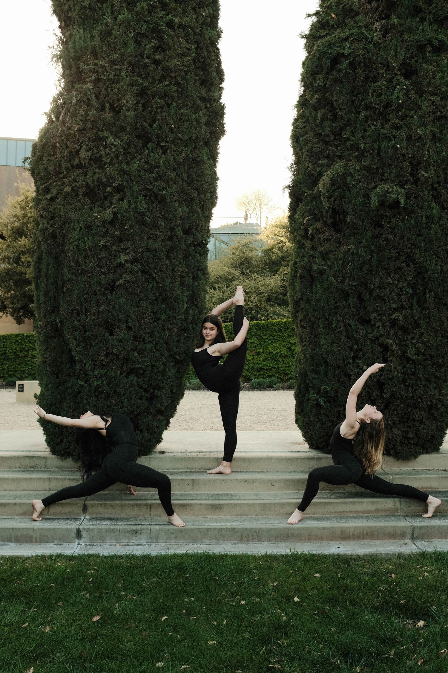 Three dancers in black attire perform coordinated poses on steps between cypress trees.