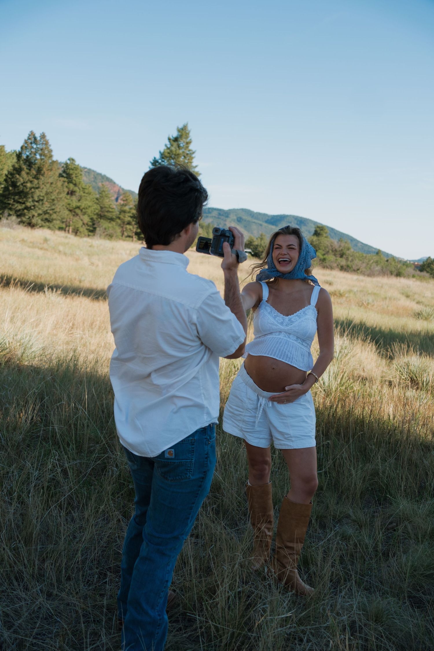 A man in a white shirt photographs a smiling pregnant woman in a blue headscarf and boots in a sunny mountain meadow.