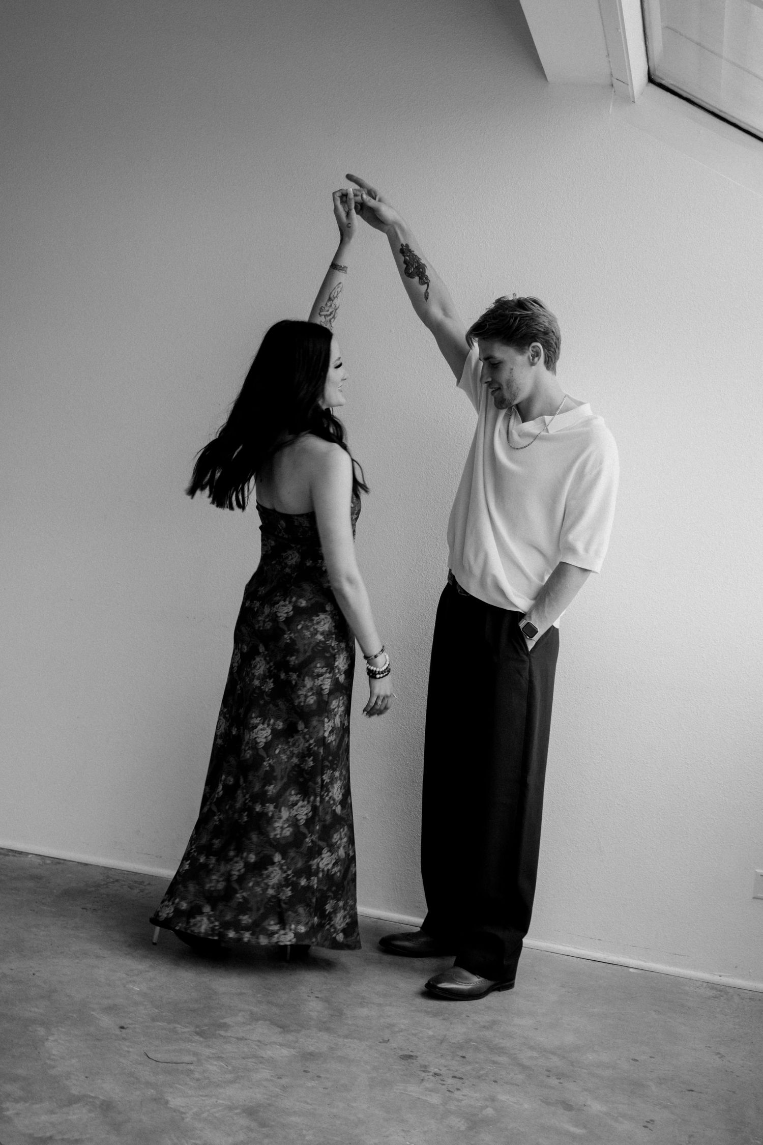 Black and white photo of a couple dancing together, man spinning woman in floral dress indoors.