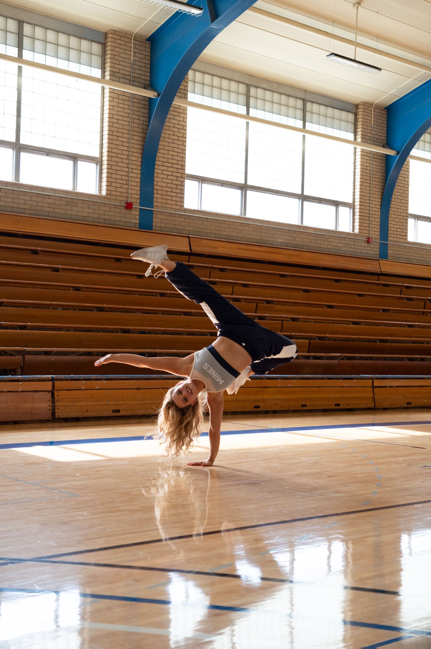 Young girl performing a one-handed cartwheel on a polished gymnasium floor with wooden bleachers in background.