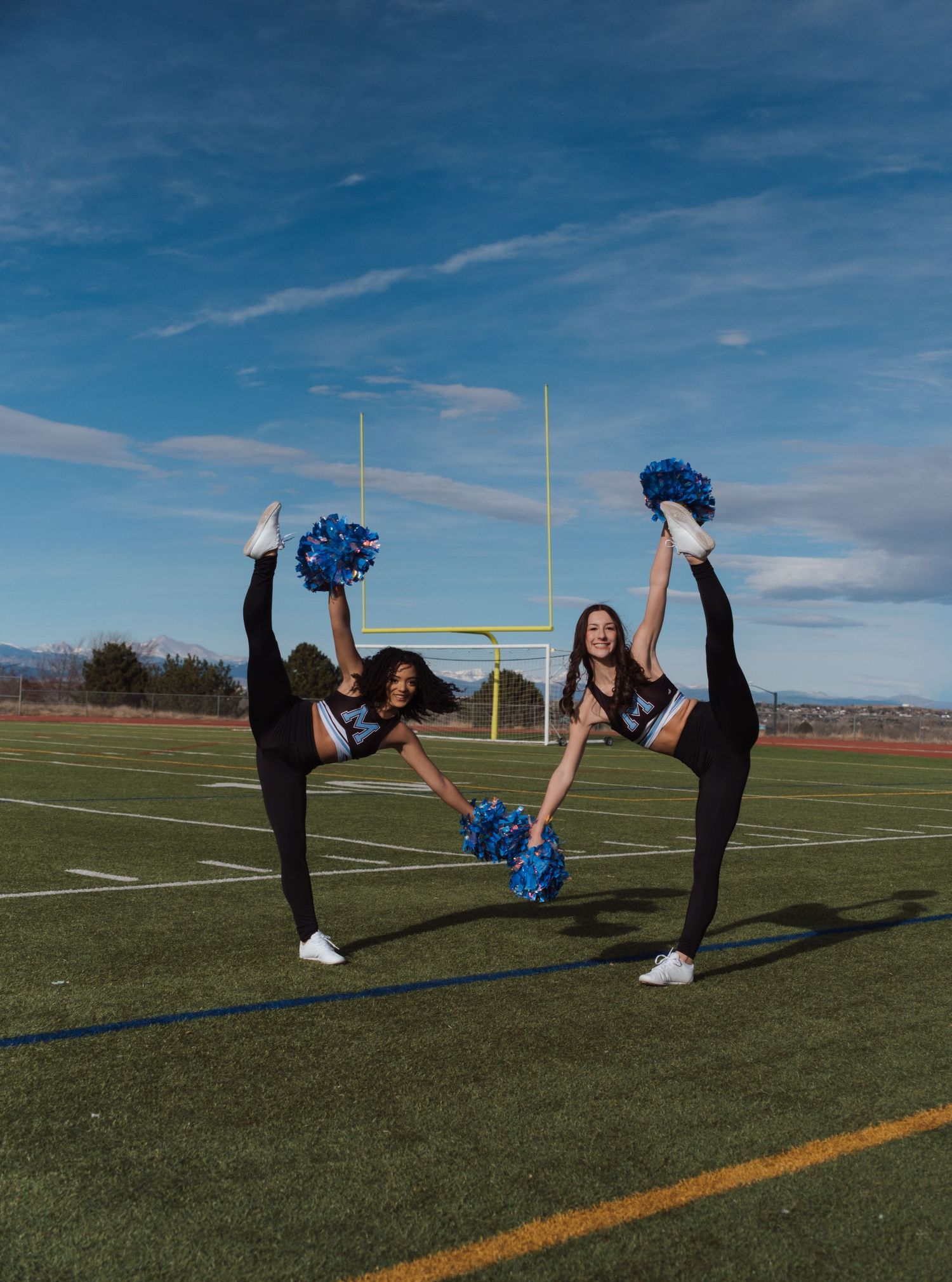 Three cheerleaders in black uniforms perform high kicks with blue pom-poms on a football field under a blue sky.